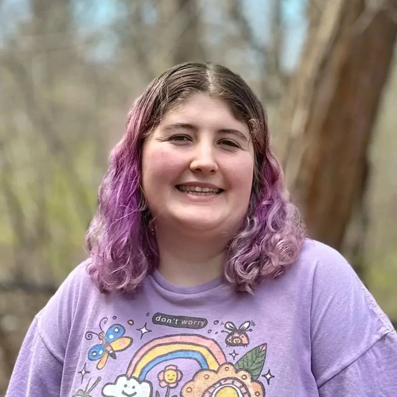 A young woman with curly purple hair smiling outdoors in a forested area, wearing a purple T-shirt with colorful cartoon designs.