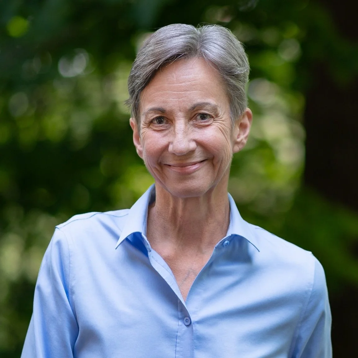 Portrait of an older woman with short gray hair, smiling, wearing a light blue button-up shirt, outdoors with a blurred green background.