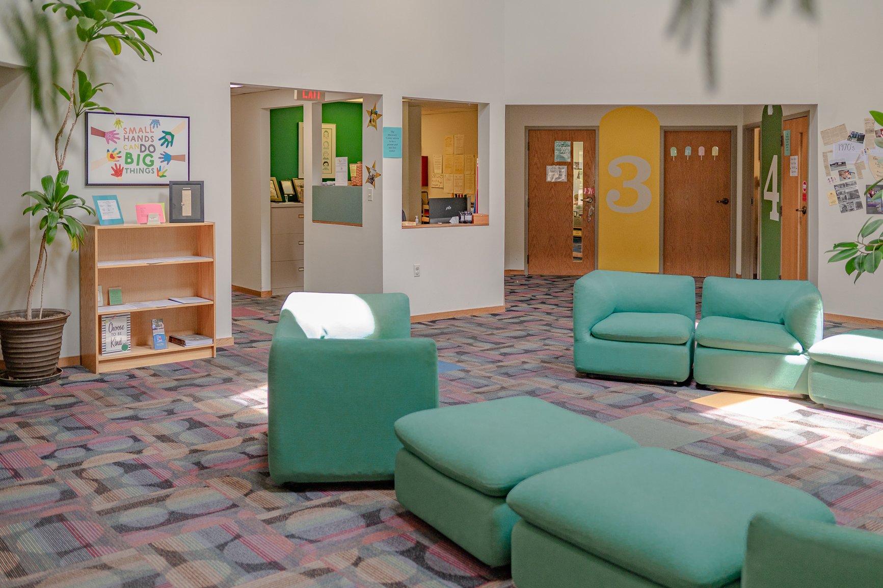Lobby area with green seating, potted plant, bookshelves, colorful art and numbered doors.