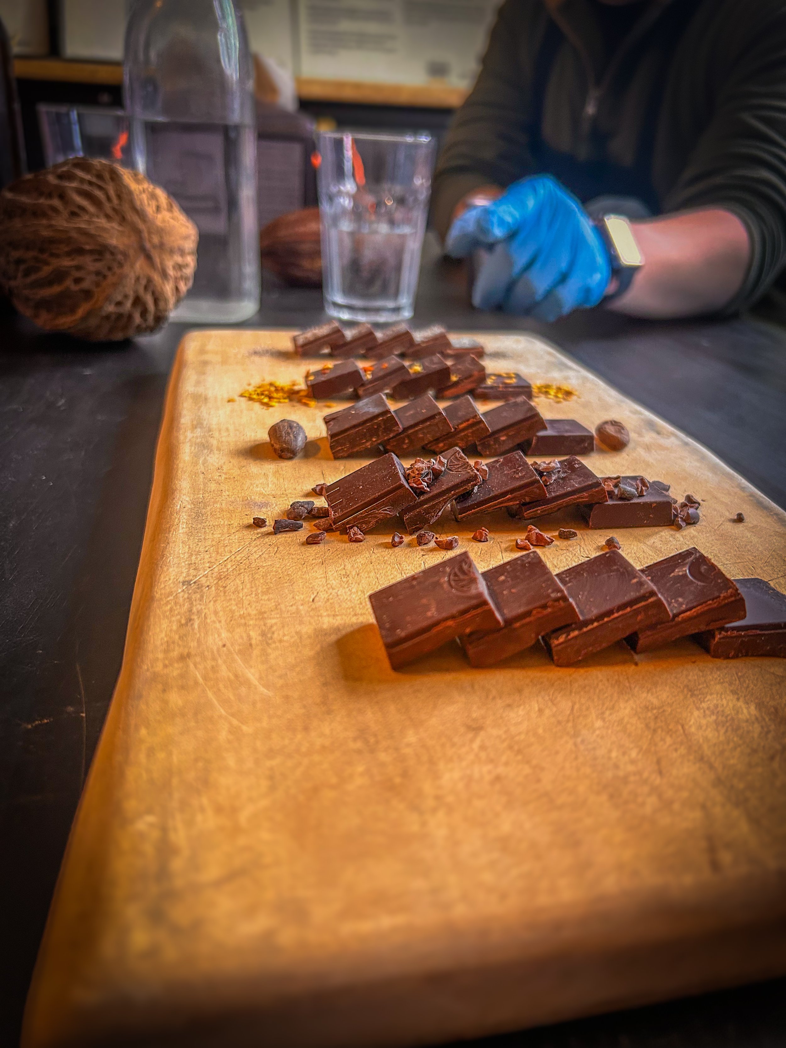 Chocolate pieces arranged on a wooden board with cacao nibs and a person's hand wearing a blue glove in the background.