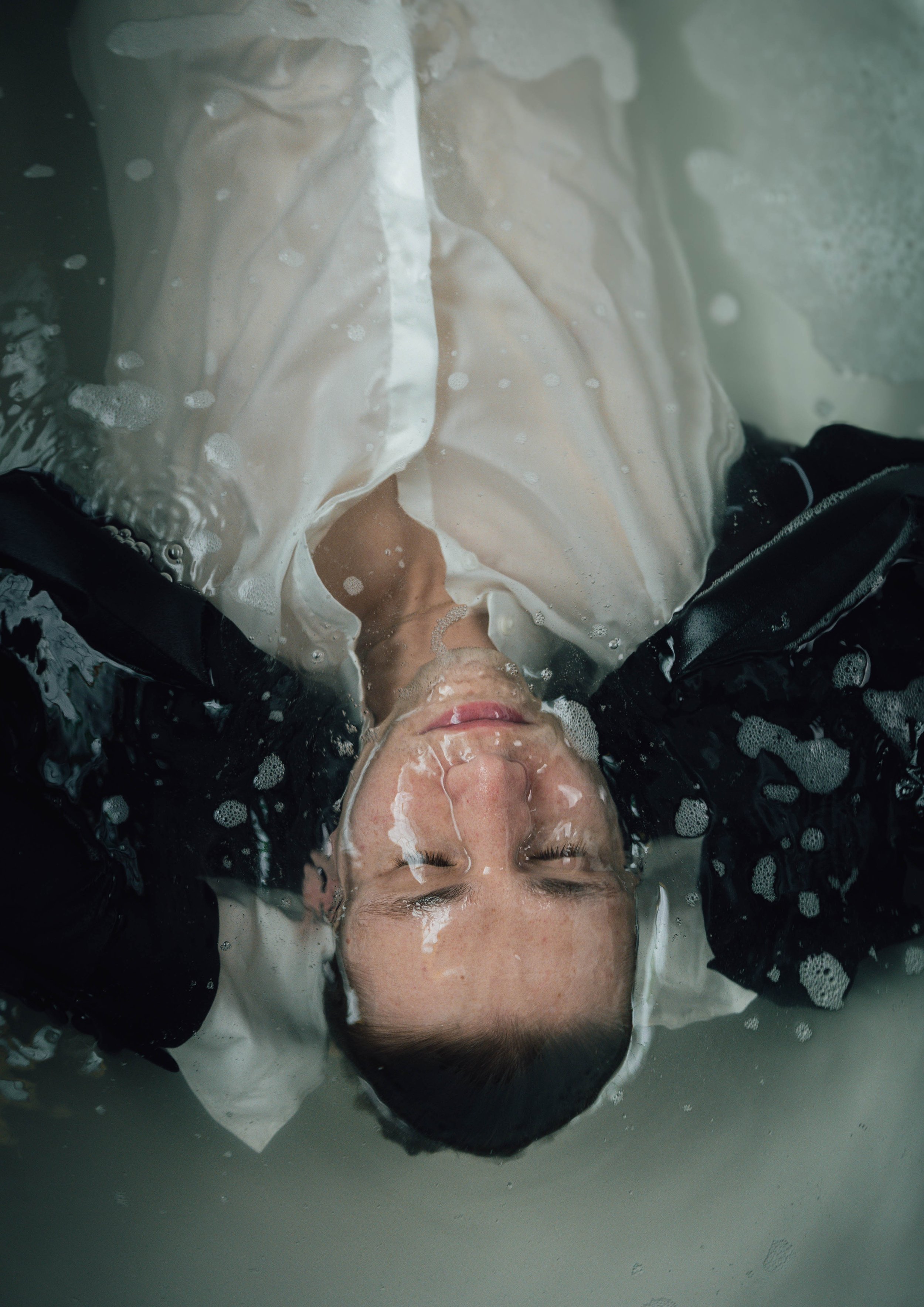 Person in water wearing a white shirt and black jacket, partially submerged with eyes closed.