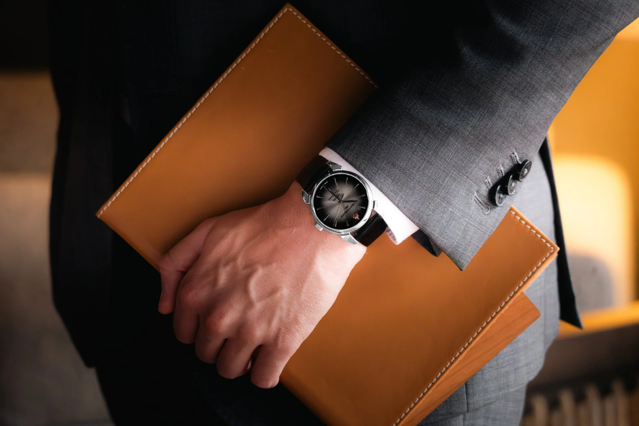 Close-up of a man in a gray suit holding a tan leather folder, wearing a silver wristwatch with a black strap.