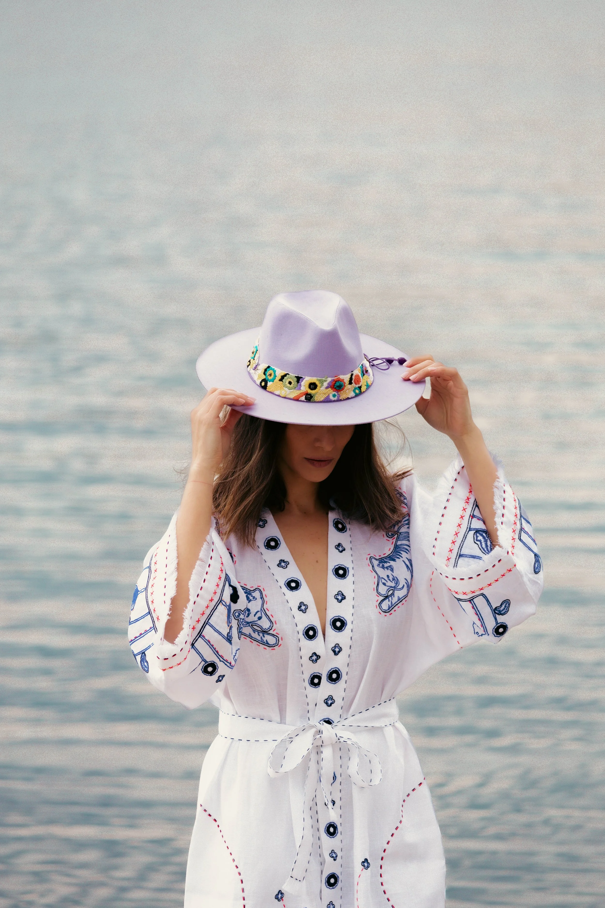 Woman in a white embroidered dress with colorful designs, wearing a lavender hat with a patterned band, standing near water.
