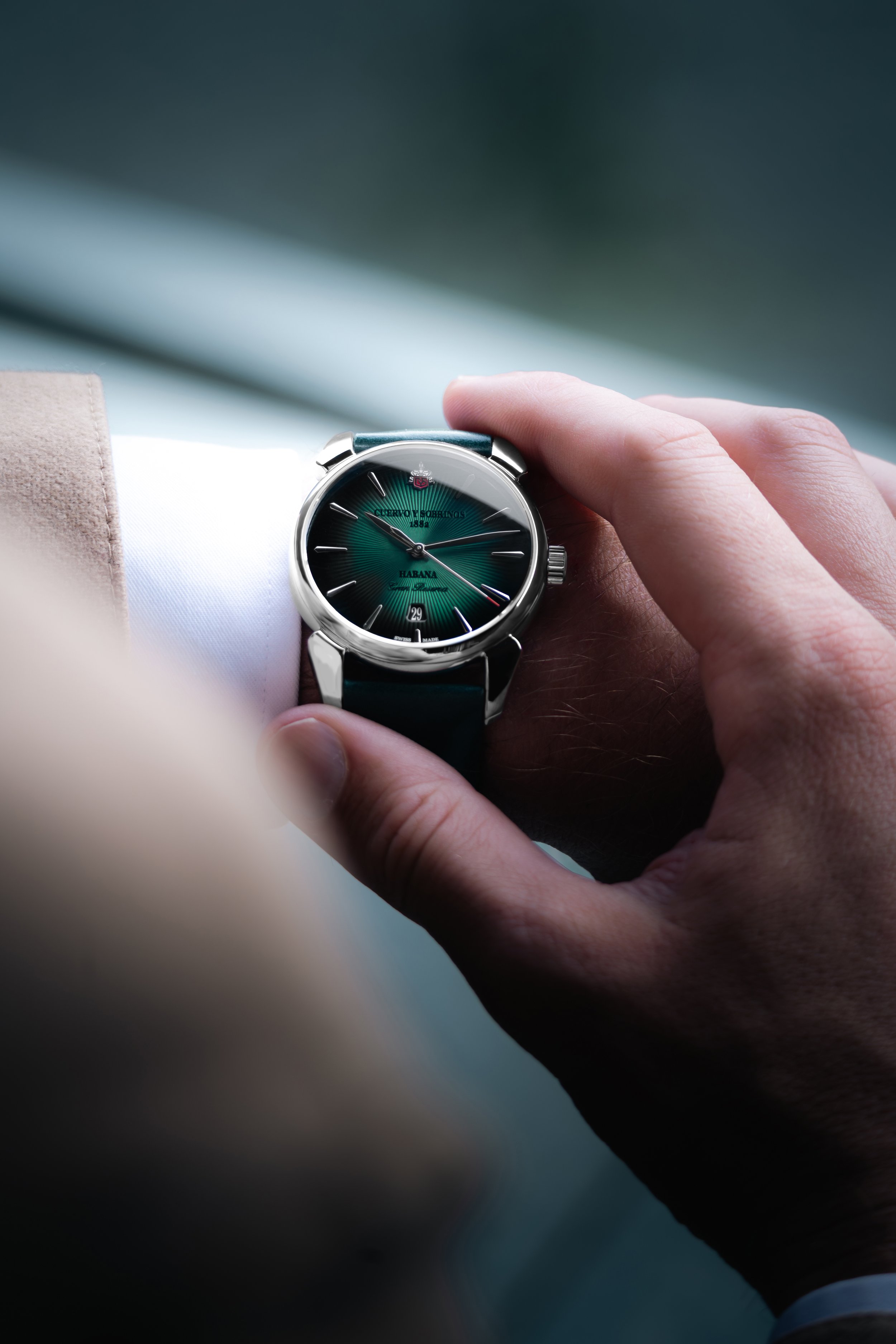 Close-up of a person's hand adjusting a luxury wristwatch with a black face and silver bezel, worn over a white shirt and beige jacket.
