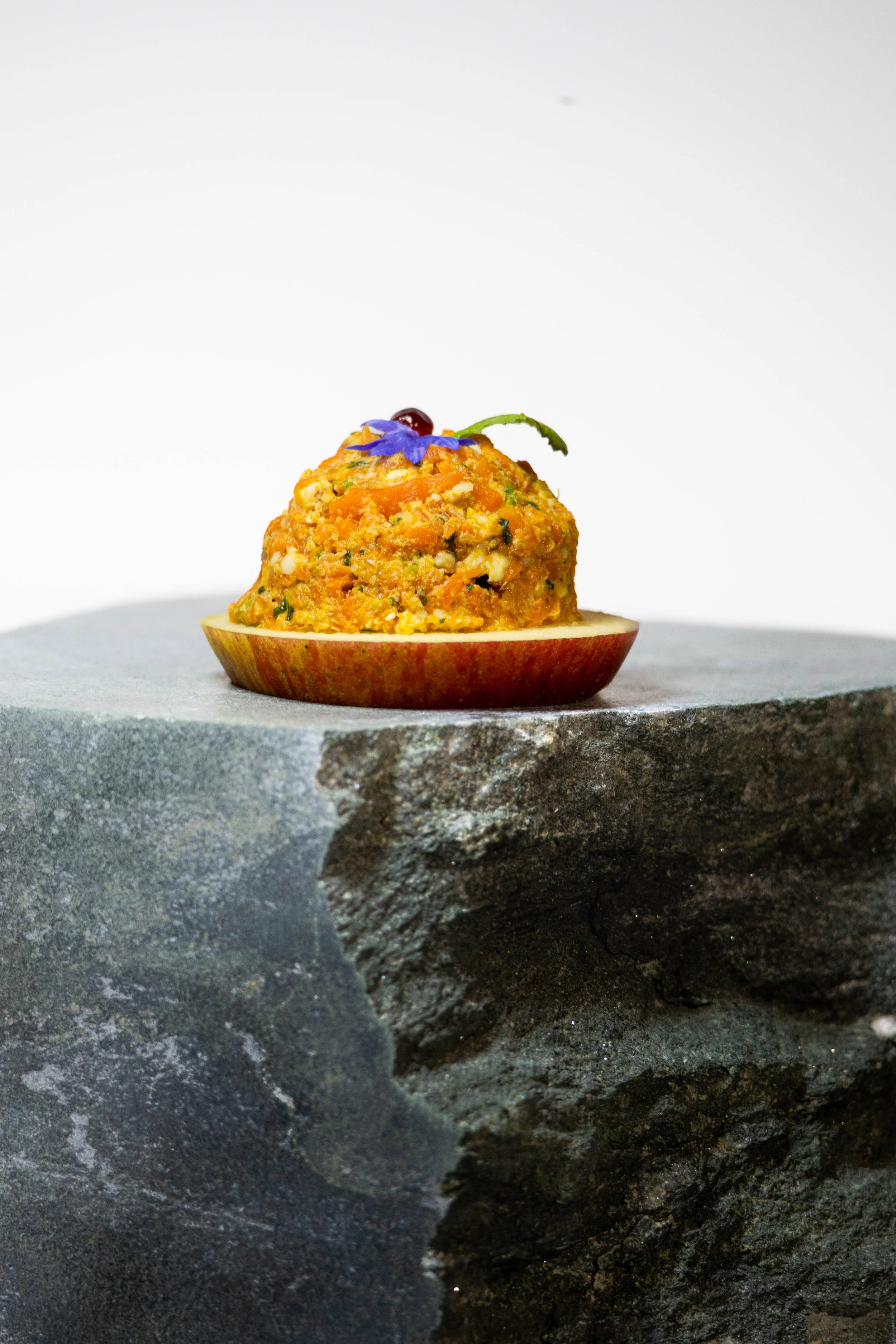A small serving of a cooked dish, possibly a vegetable or grain mixture, is in a halved apple shell, garnished with a purple edible flower, atop a gray stone surface.