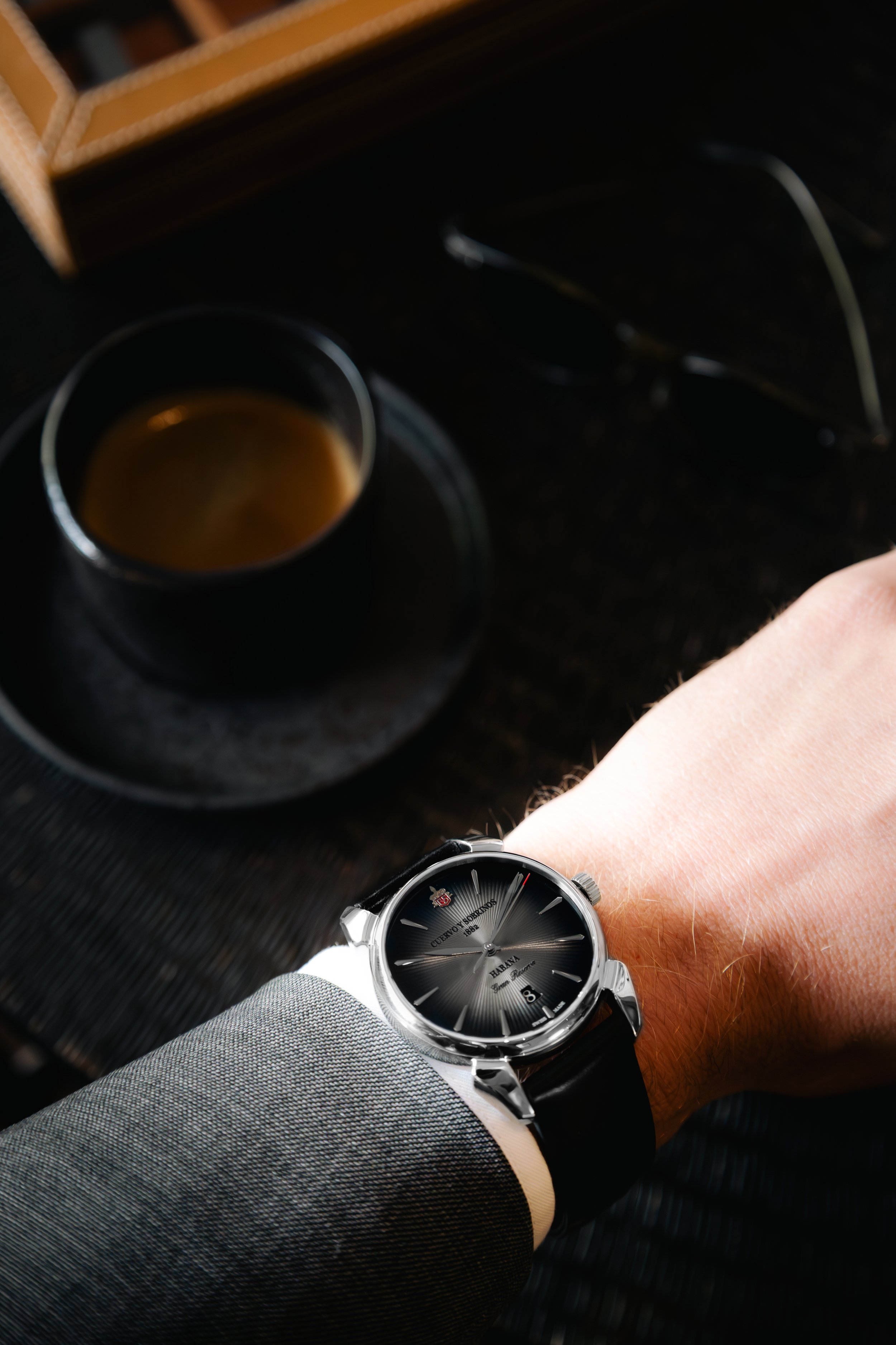 A person's wrist wearing a black leather strap watch with a black dial, silver case, and a business suit sleeve, next to a cup of coffee on a black textured surface, with glasses nearby.