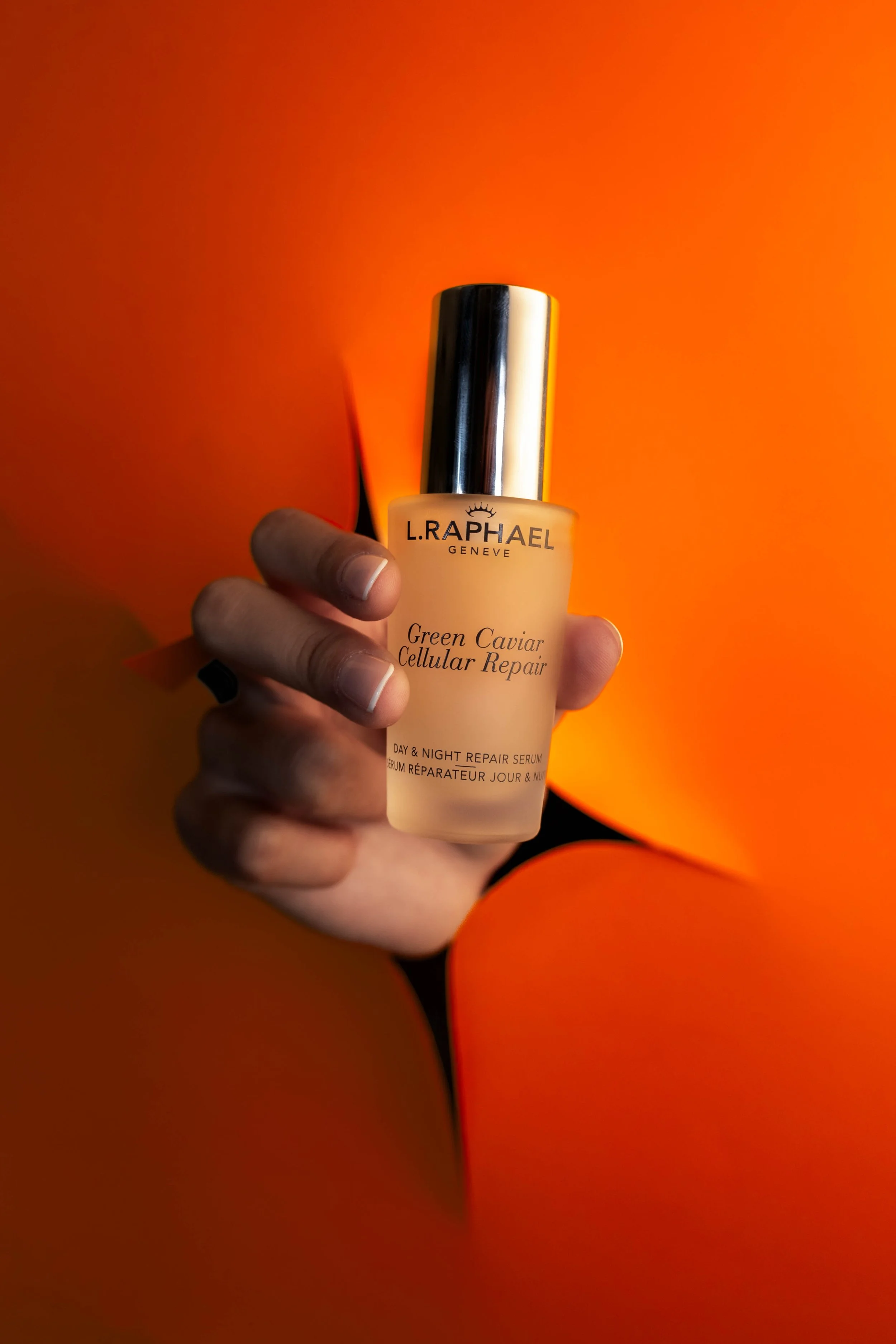 Hand holding L. Raphael skincare serum against orange background.