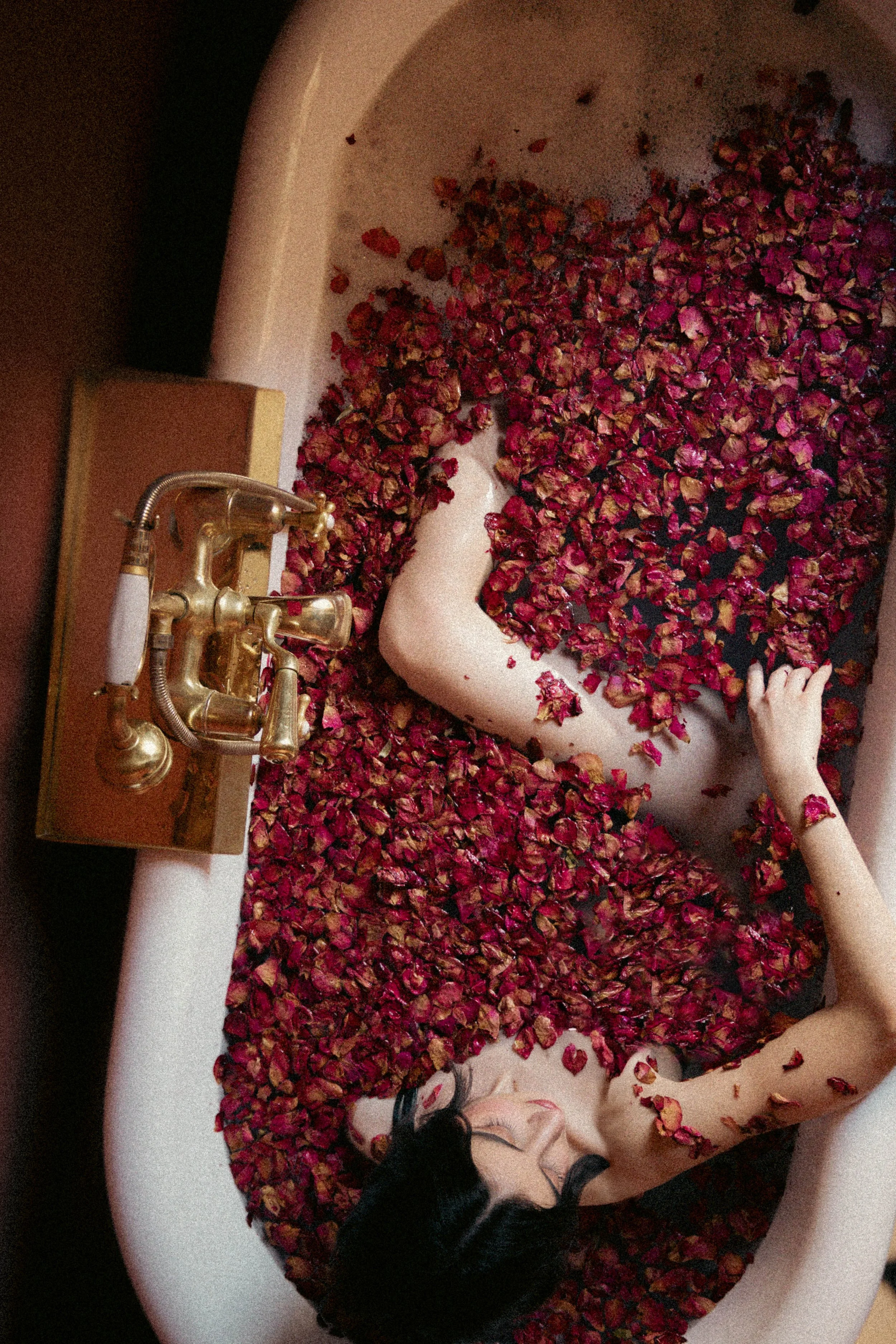 Woman in bathtub filled with red rose petals and water, vintage brass faucet.