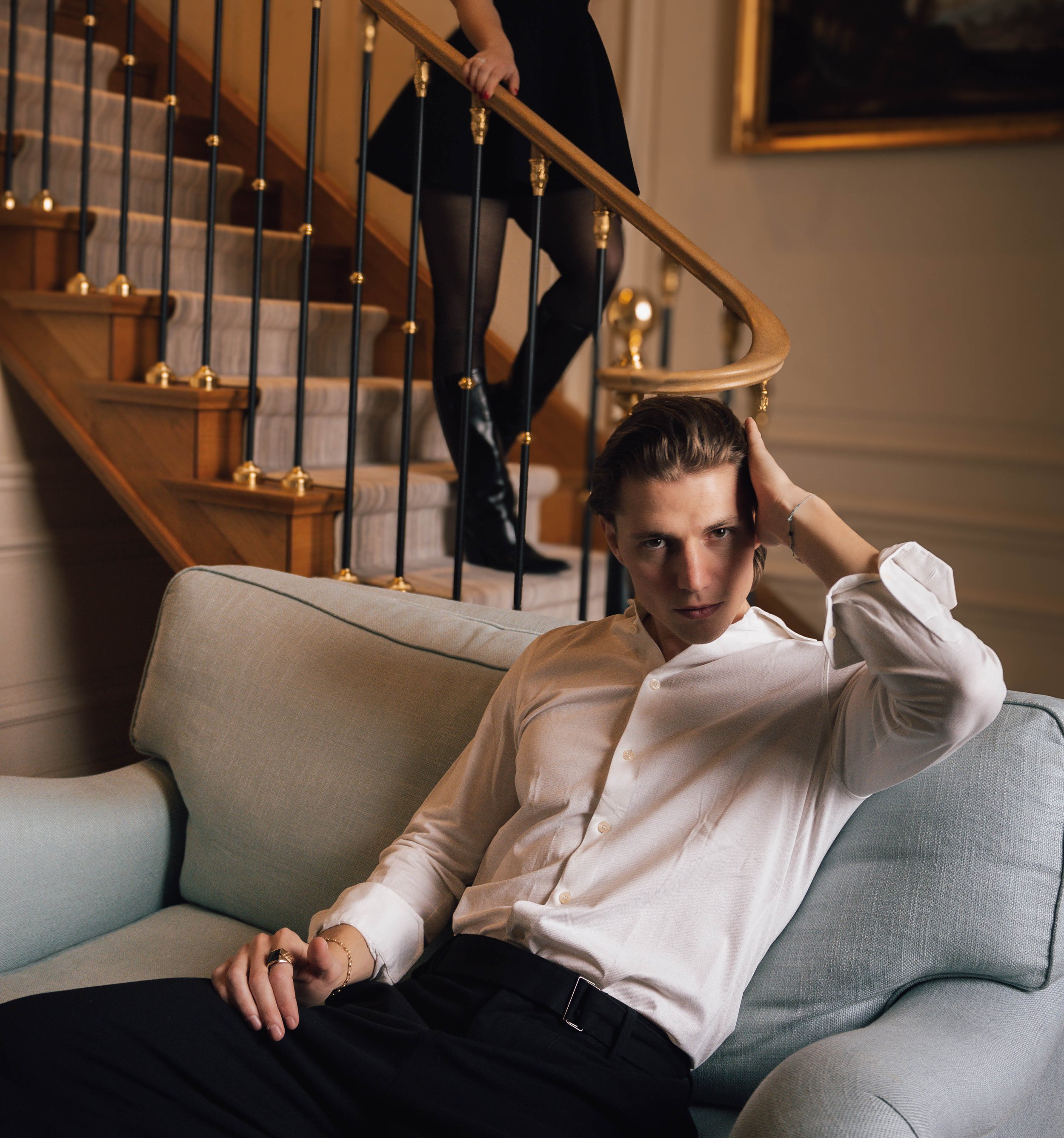 A person in a white shirt sitting on a light blue sofa in front of a staircase, while another person's legs are visible on the stairs.