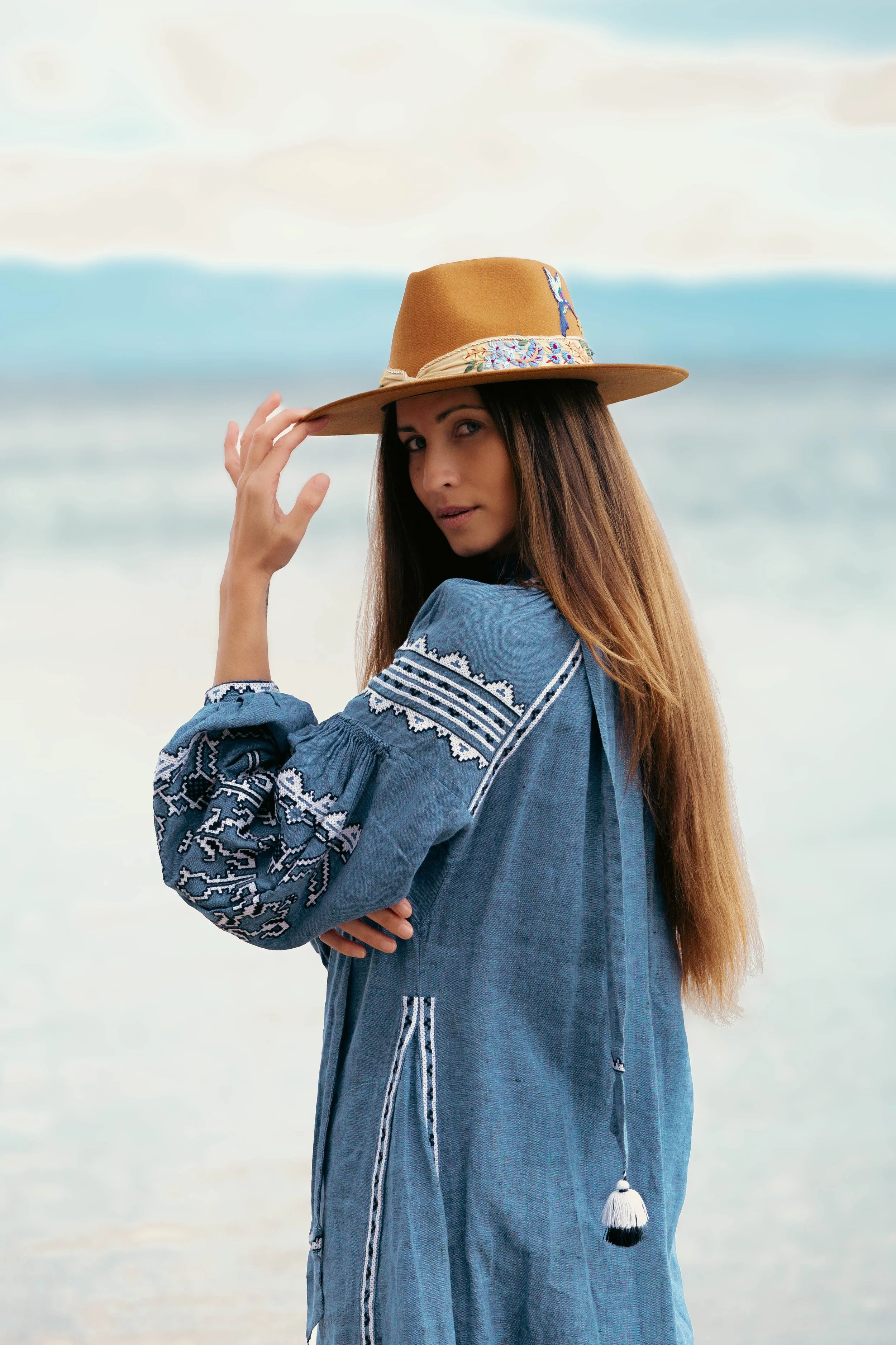 Woman in a blue embroidered dress and brown hat by the sea