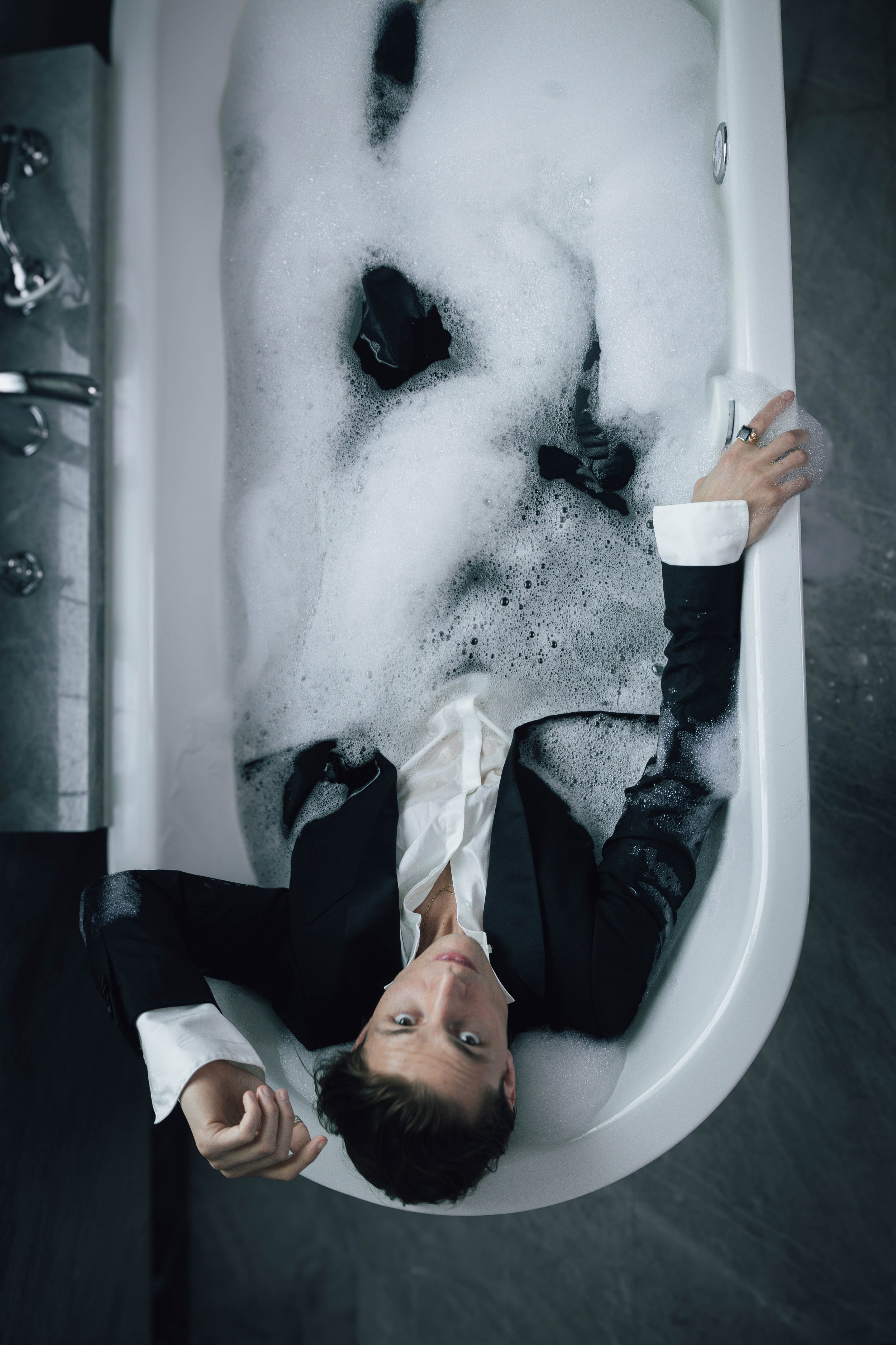 Person in a suit lying in a bathtub filled with bubbles.