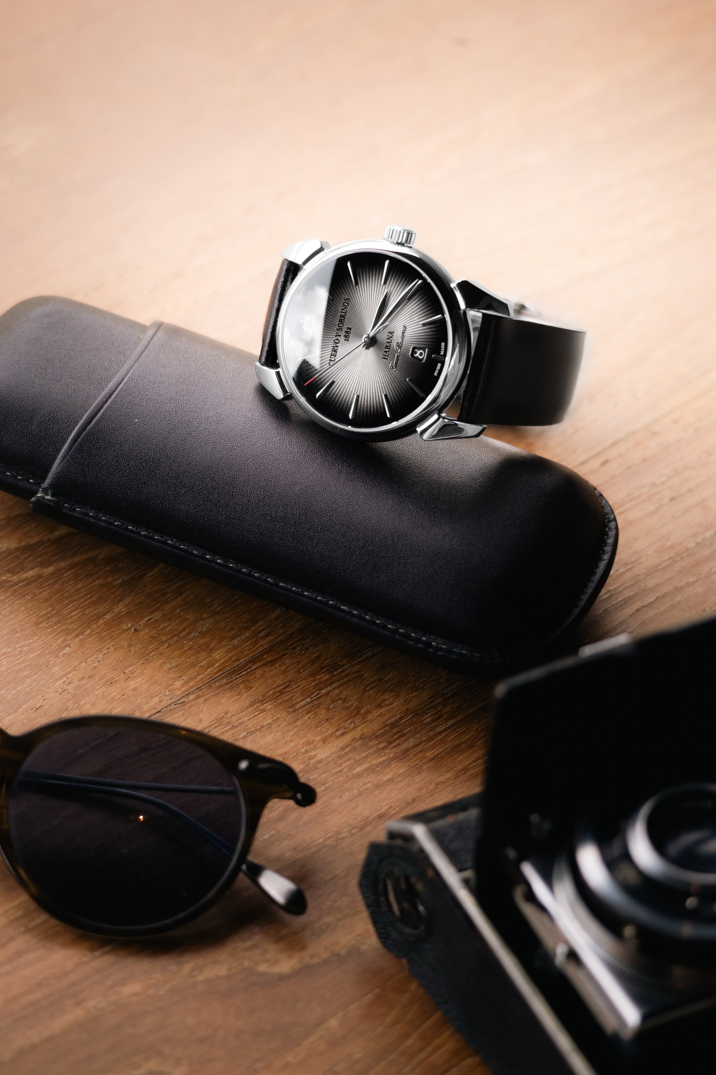 A black wristwatch with a silver case on a black leather strap, resting on a black glasses case on a wooden surface. Part of a camera and a pair of sunglasses are also visible.