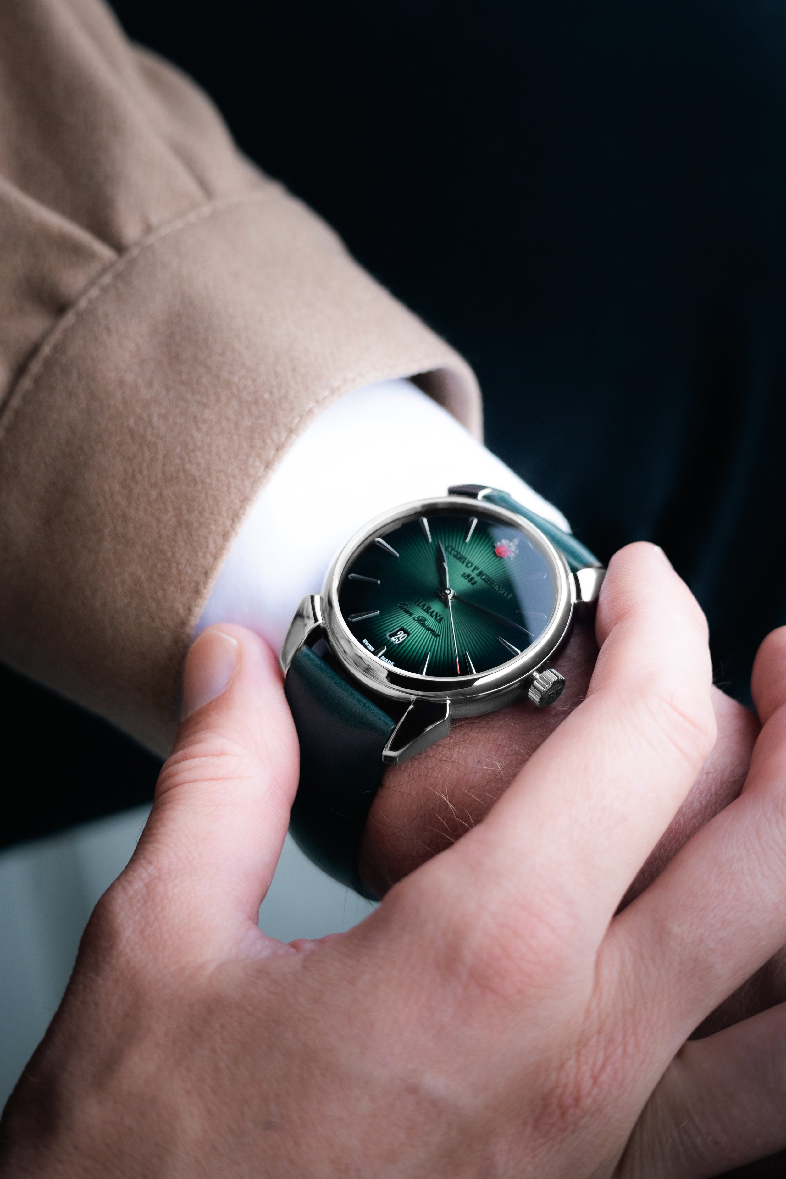 A person wearing a beige coat and white shirt adjusting a wristwatch with a green dial and black strap.