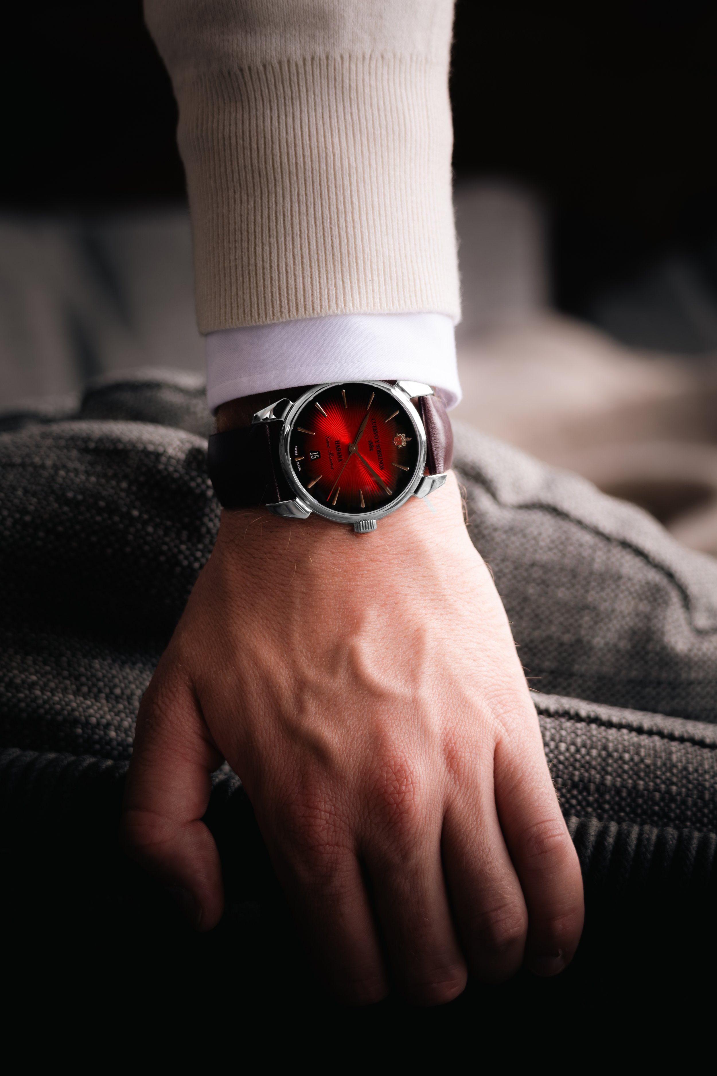 A person's hand wearing a wristwatch with a red dial, black leather strap, and metal case, resting on gray fabric.