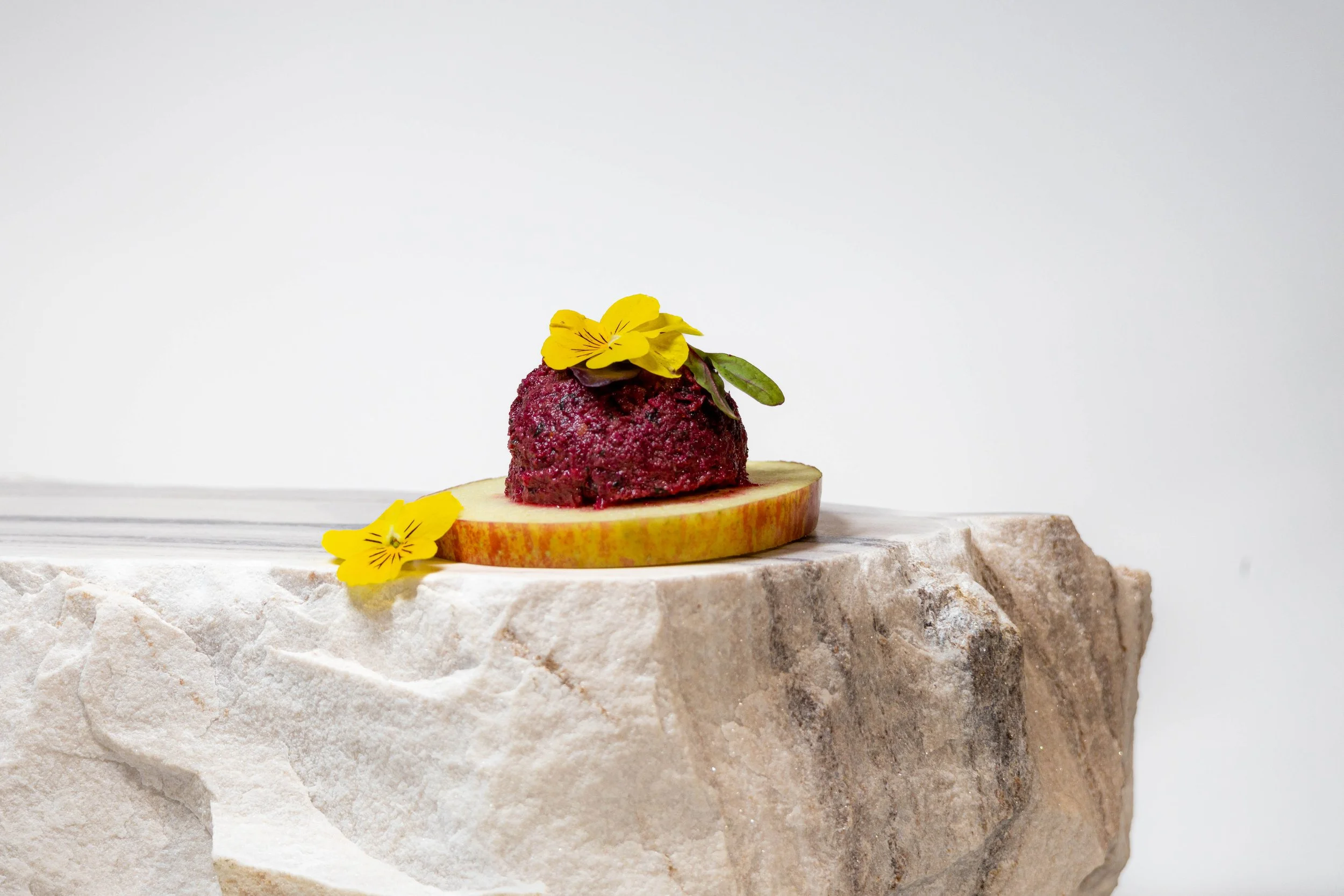 A small, round red scoop of what appears to be sorbet or ice cream on a slice of apple with a yellow flower on top and a yellow flower beside it, all displayed on a textured stone surface against a plain white background.