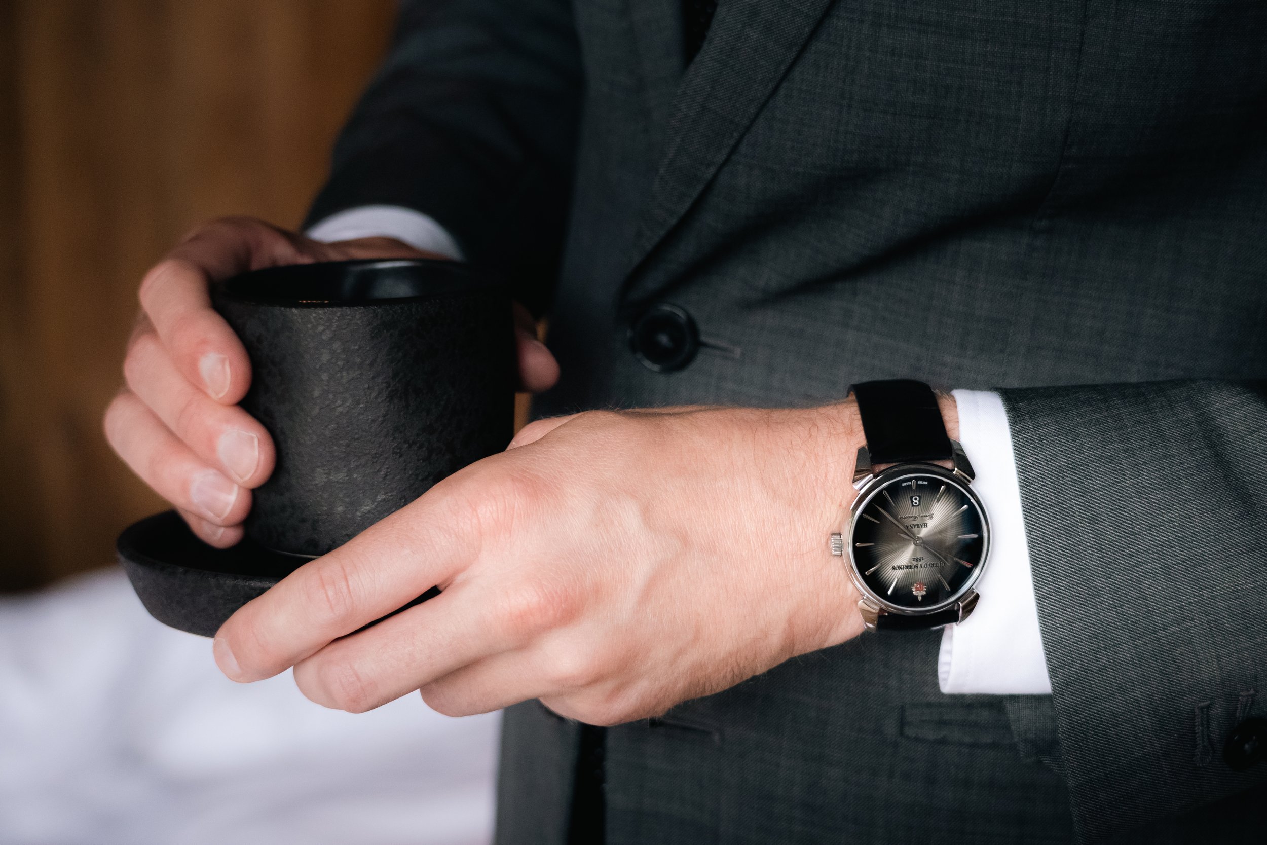 A man in a gray suit holding a black mug, wearing a silver watch with a black leather strap.
