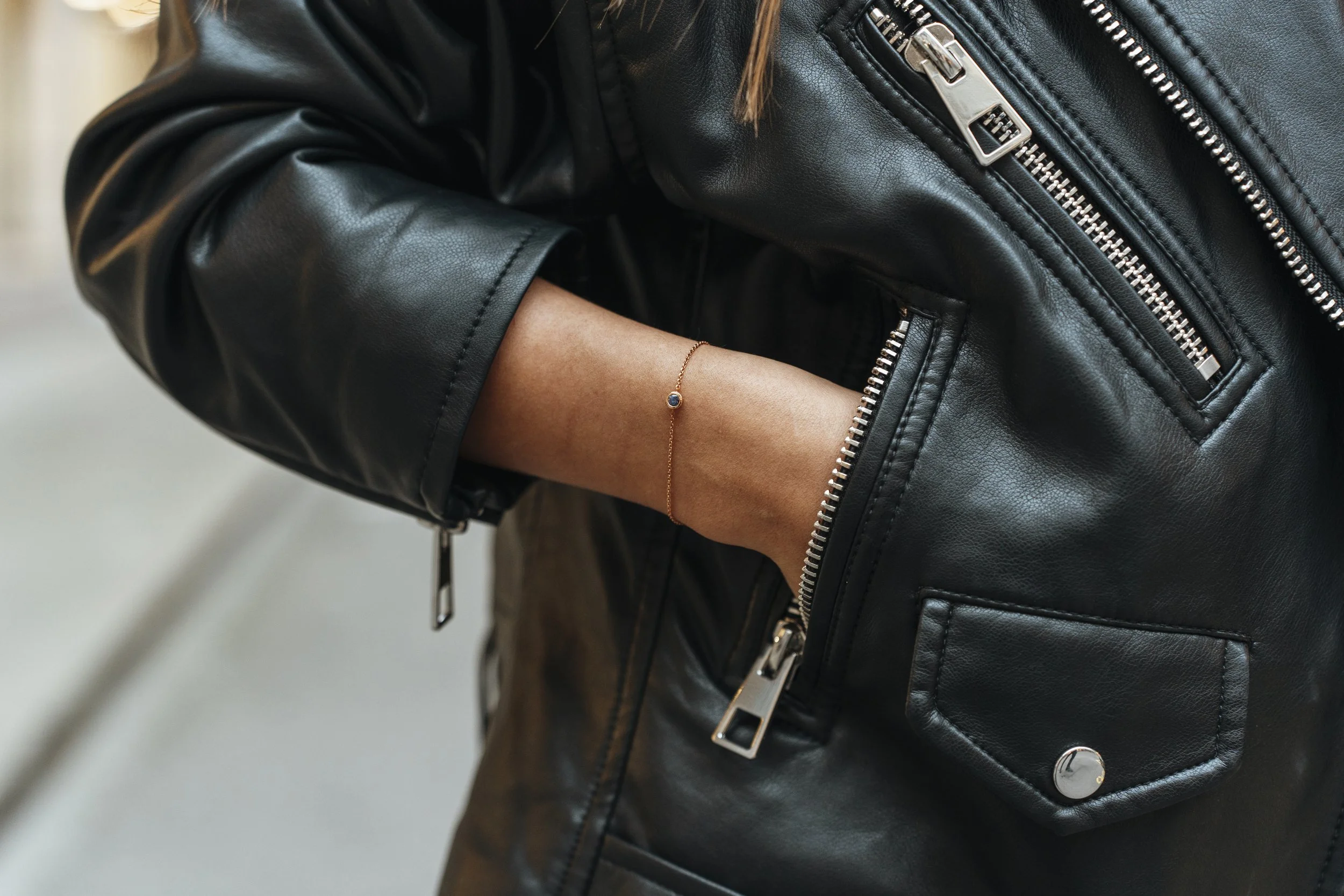 Close-up of a person wearing a black leather jacket with silver zippers, hand in pocket, wearing a delicate bracelet.