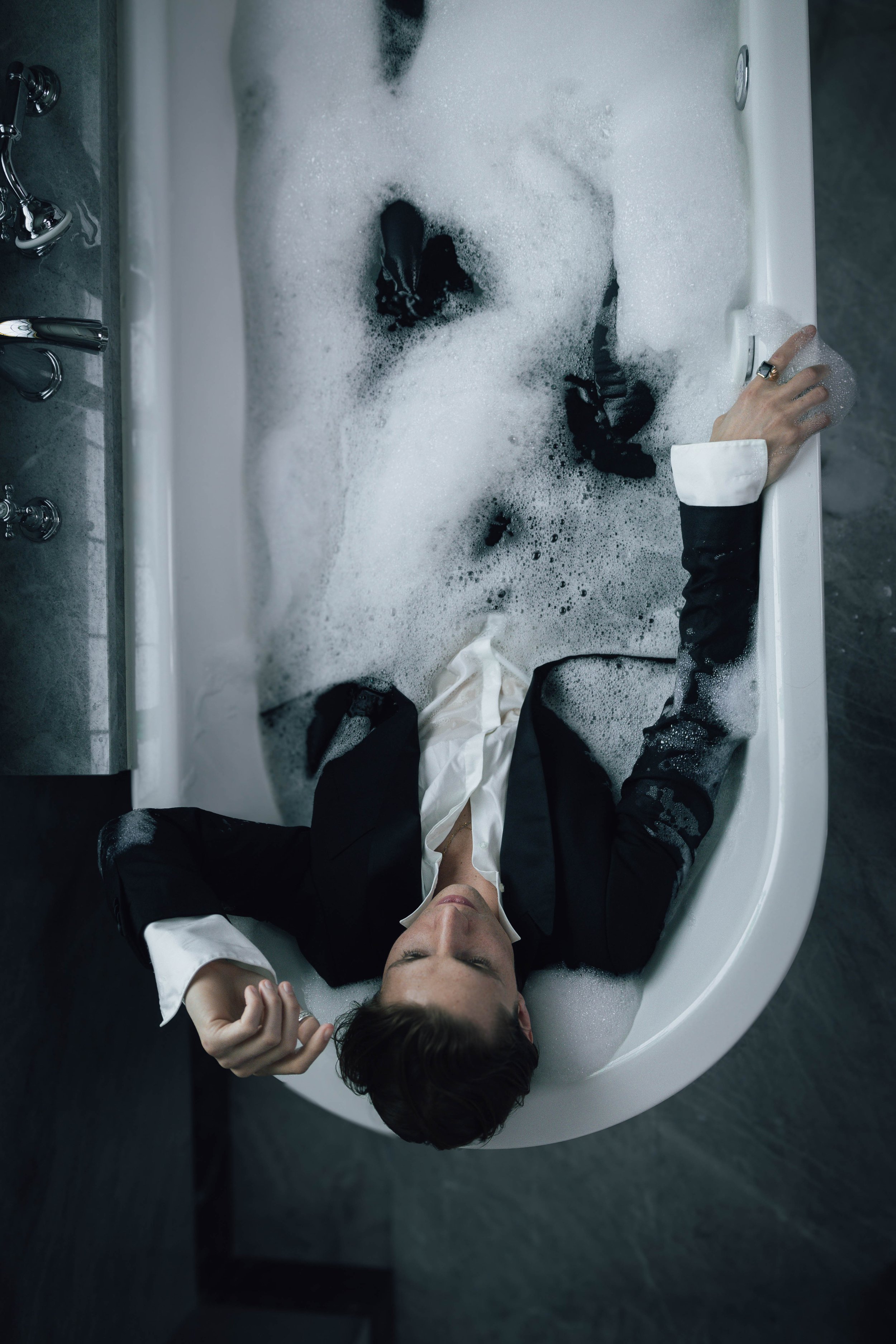 Person wearing a suit and lying in a bathtub filled with soapy water, eyes closed.