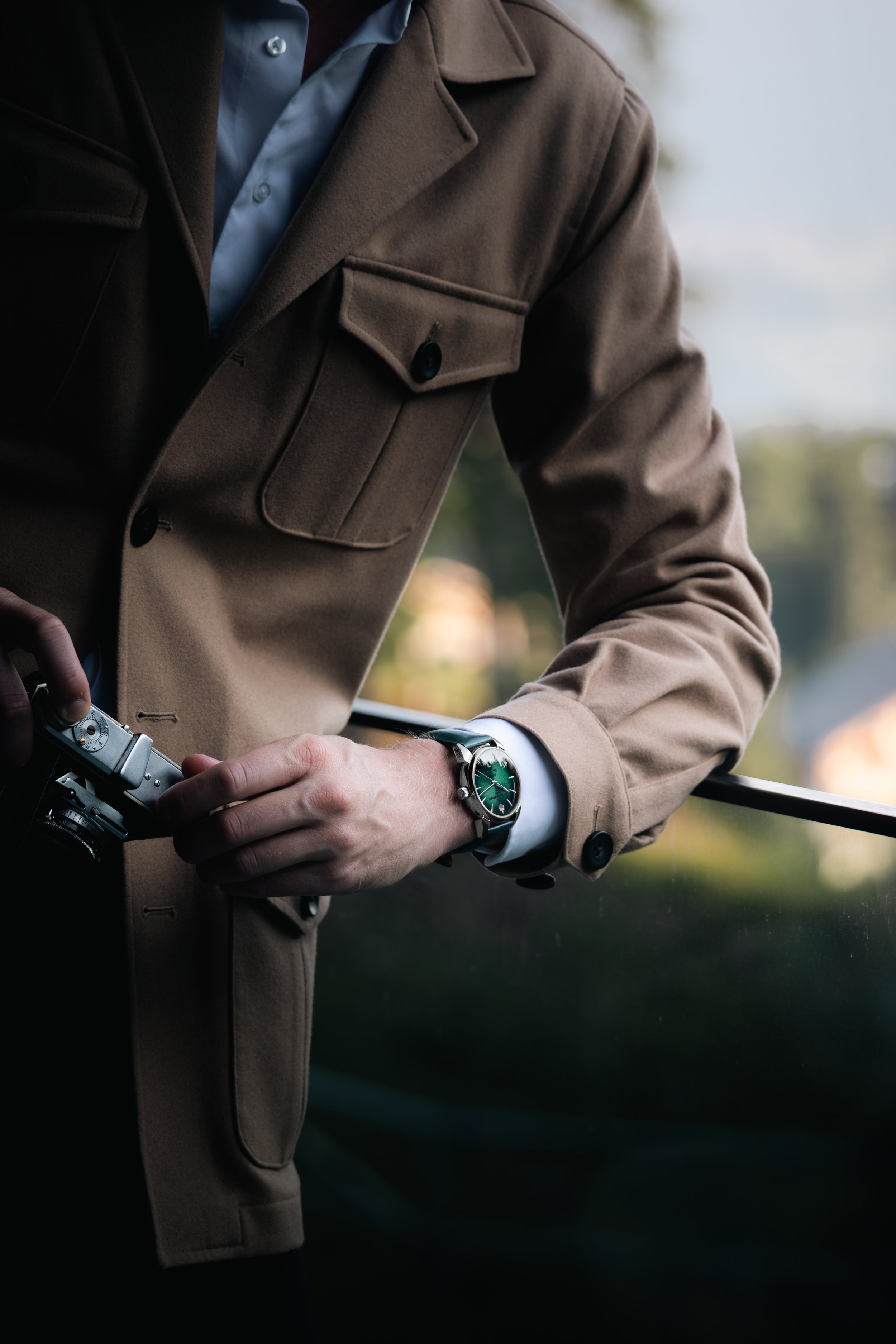 Close-up of a man wearing a tan coat and a silver wristwatch, holding a camera, standing outdoors near a railing with a blurred nature background.