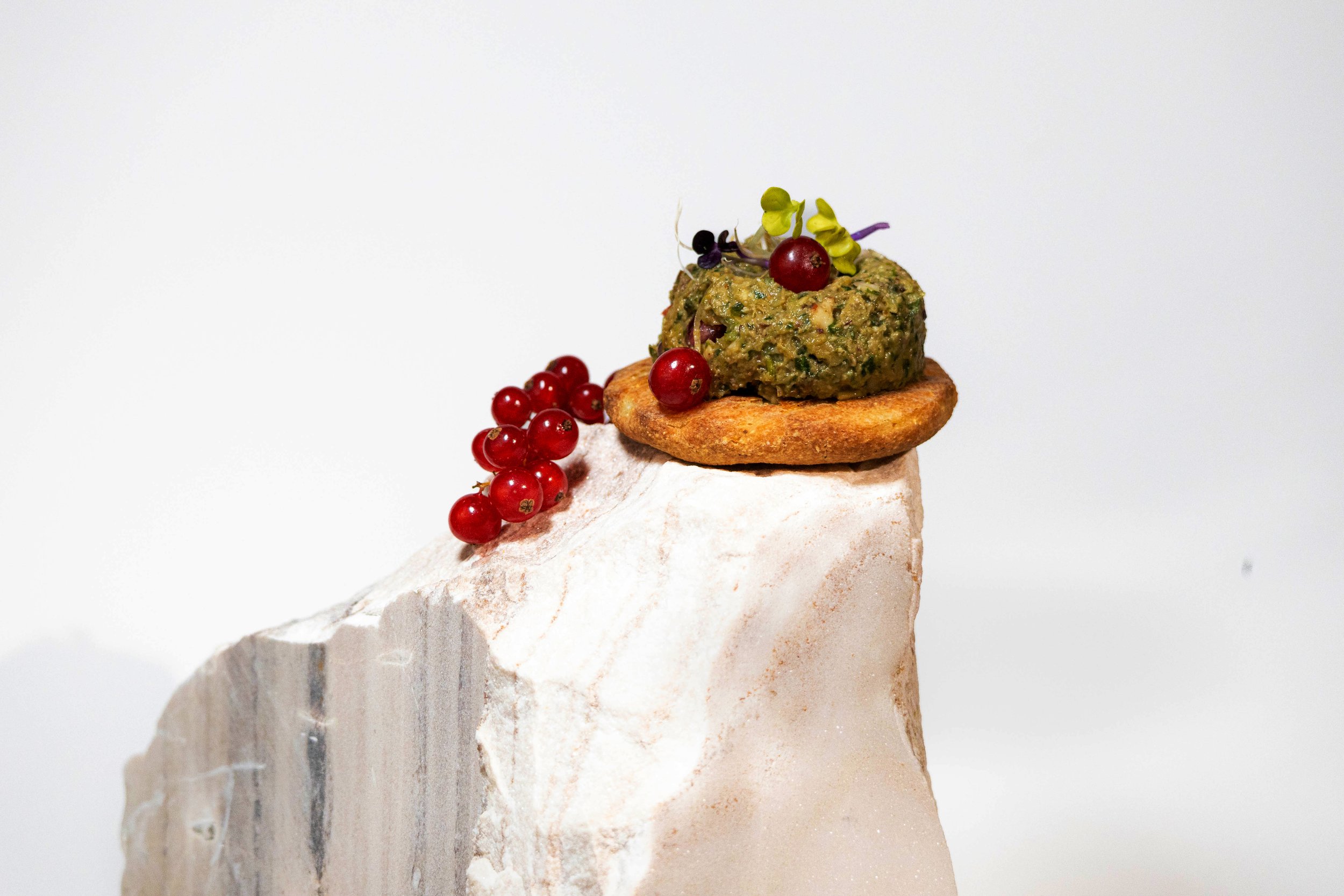 A gourmet appetizer featuring a round croquette topped with a green herb spread, garnished with microgreens and red currants, placed on a toasted round cracker, with a small cluster of red currants beside a white mineral rock.