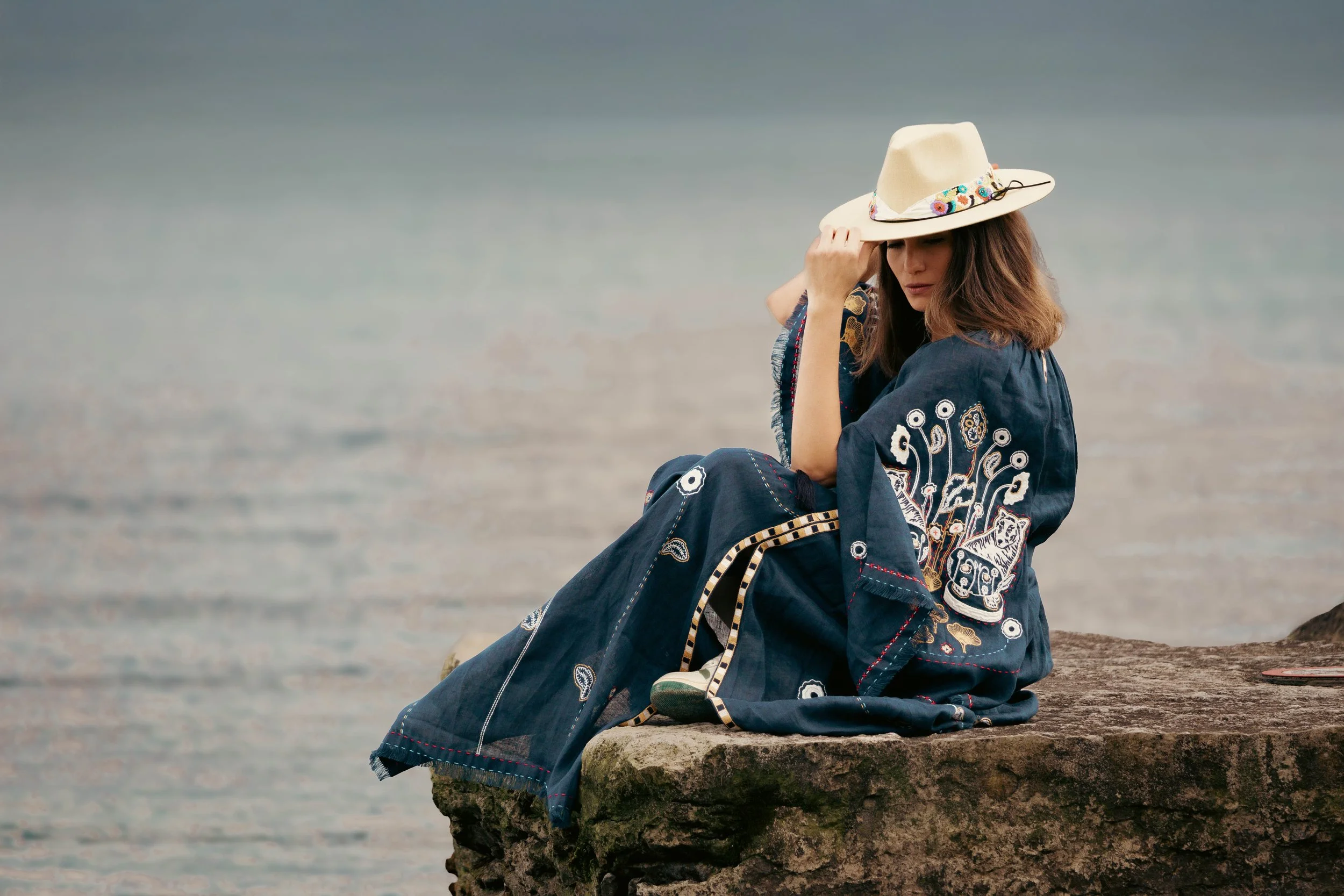 Woman wearing a decorated robe and a hat, sitting on a rock by water.