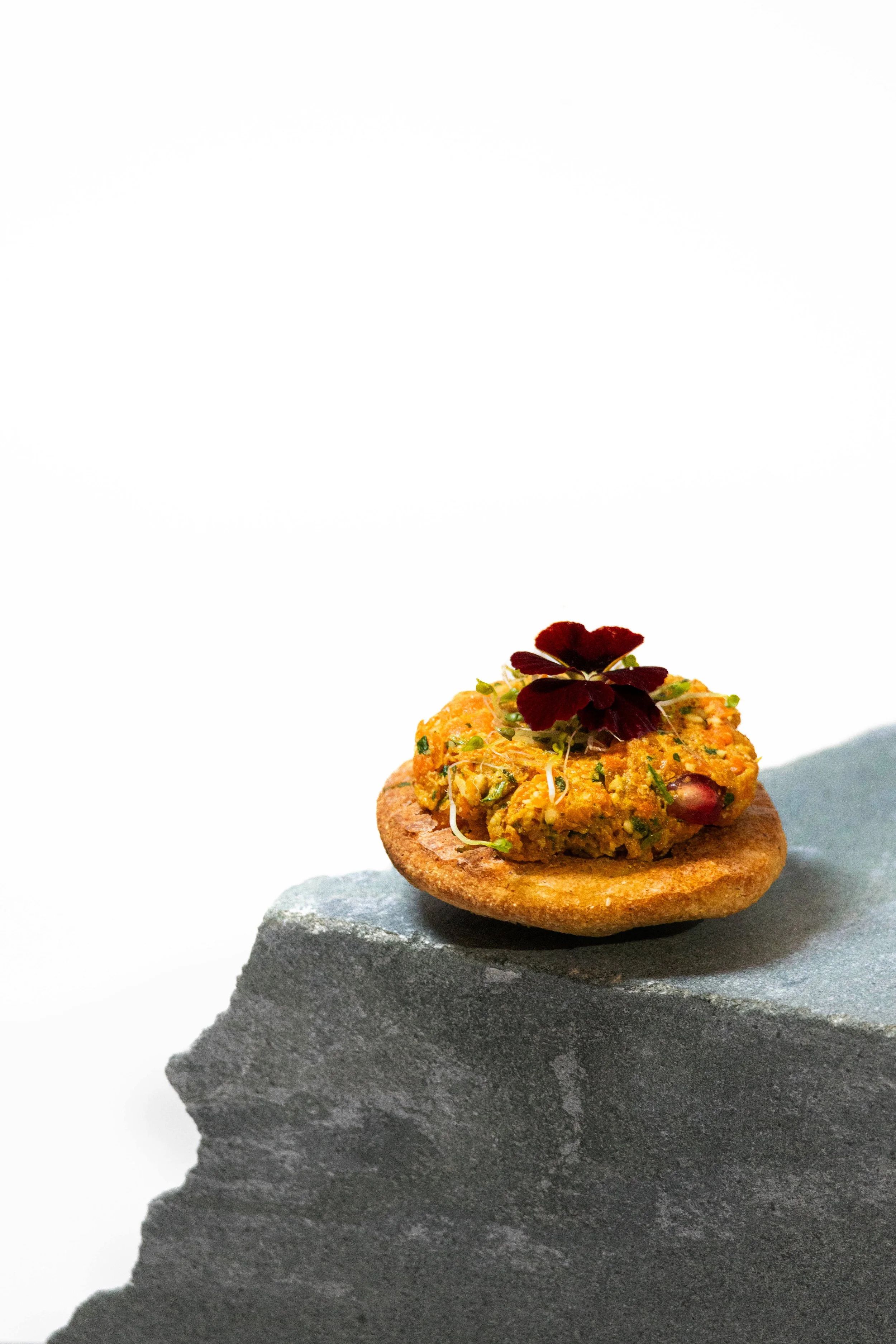 Close-up of a small round cracker topped with a dollop of colorful spread, garnished with a small dark red edible flower, placed on a gray stone surface.