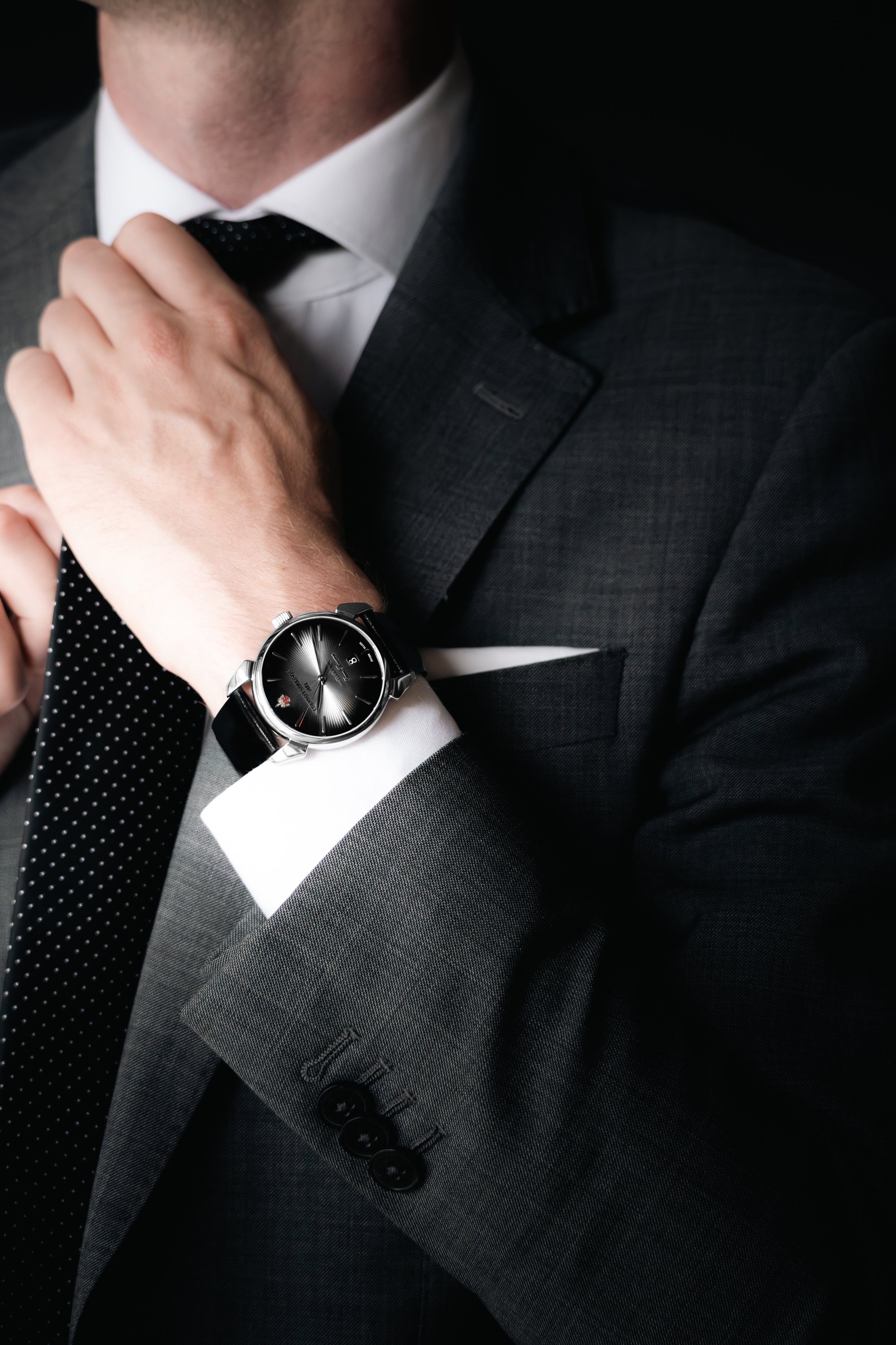 Close-up of a man in a dark suit adjusting his black polka dot tie, wearing a silver wristwatch with a black leather band on his left wrist.