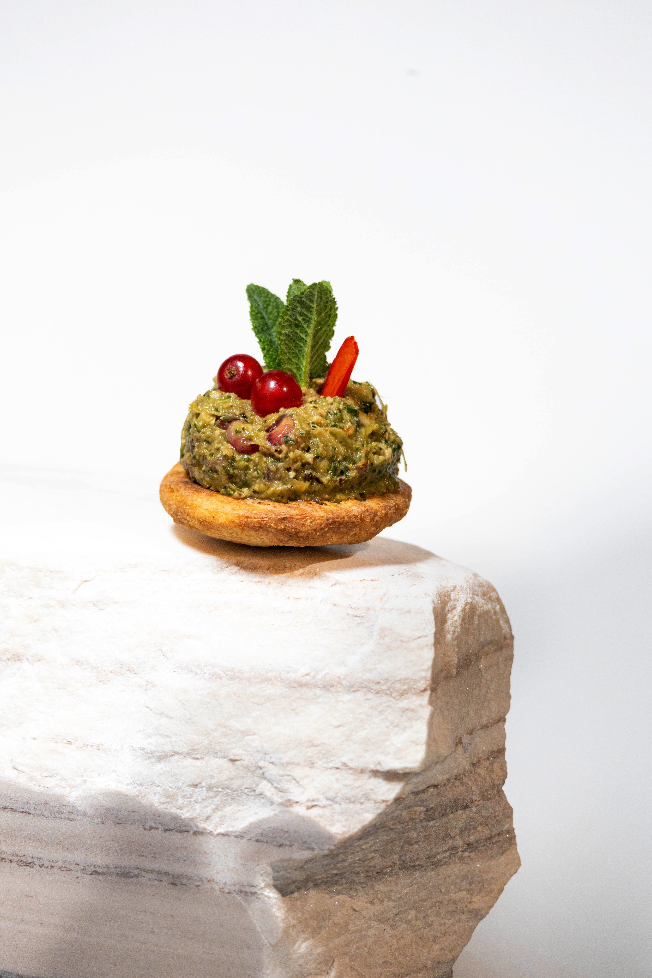 Small appetizer with green spread, red berries, a mint leaf, and red pepper on a round cracker, placed on a white rock with a plain white background.