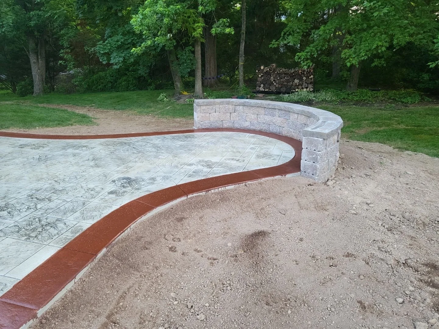 Stamped concrete patio with red-tinted border and curved stone seating wall, mid-construction