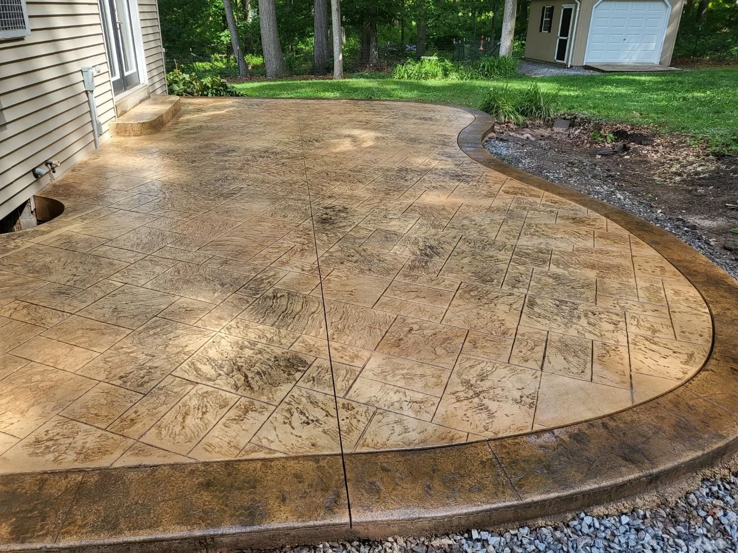 Curved tan stamped concrete patio with matching steps, next to a backyard storage shed