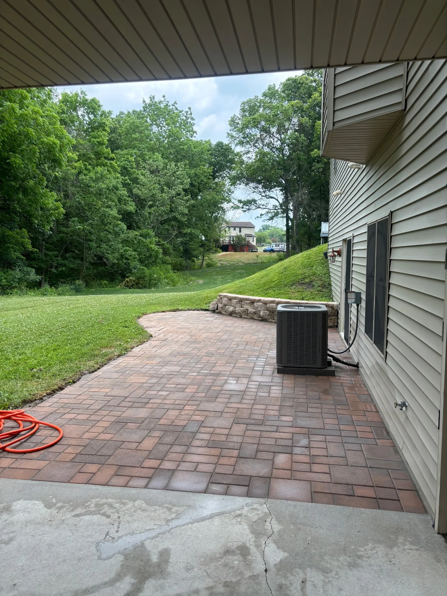 Red paver patio with a low stone retaining wall behind a home, adjacent to outdoor equipment pad