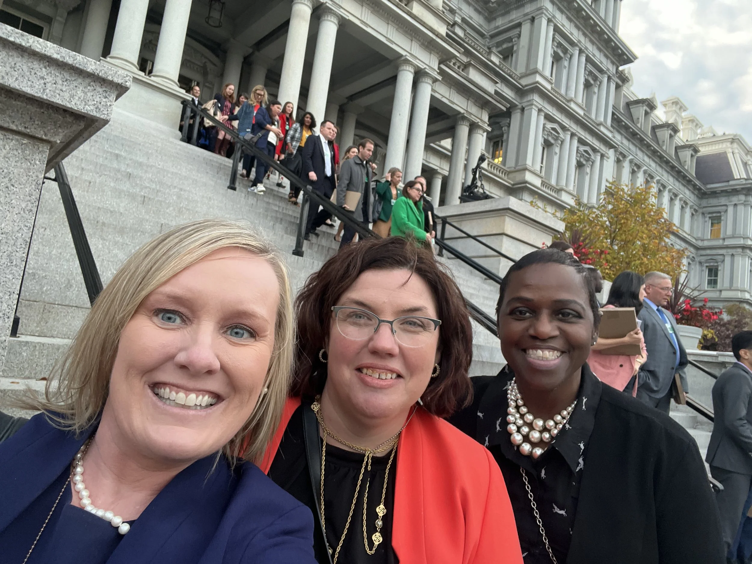 Three women smiling on the steps of a government building during a formal event, with many people in the background lining the stairs.