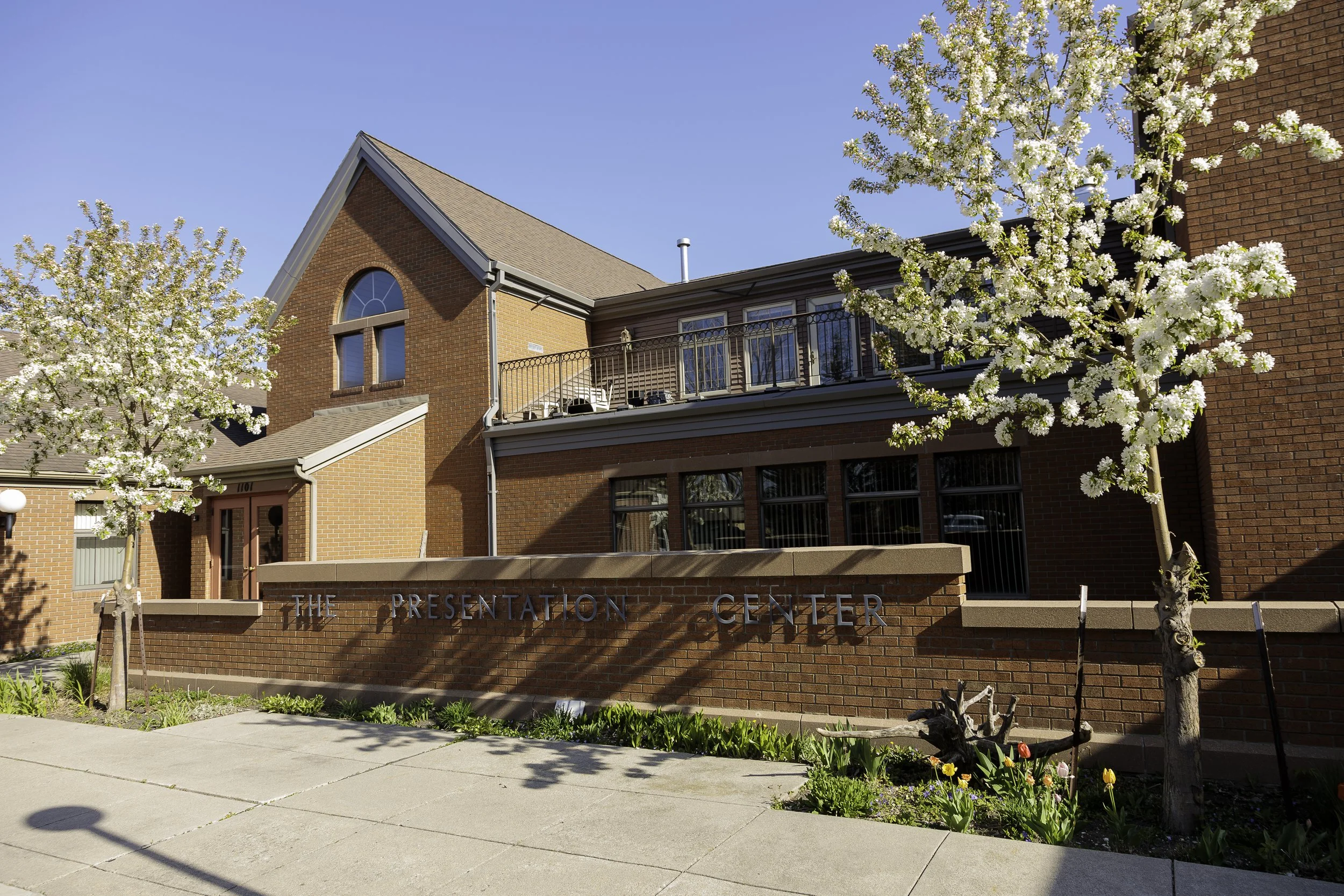 Brick building with sign reading "The Presentation Center," trees with white blossoms, a sidewalk, and a clear blue sky.