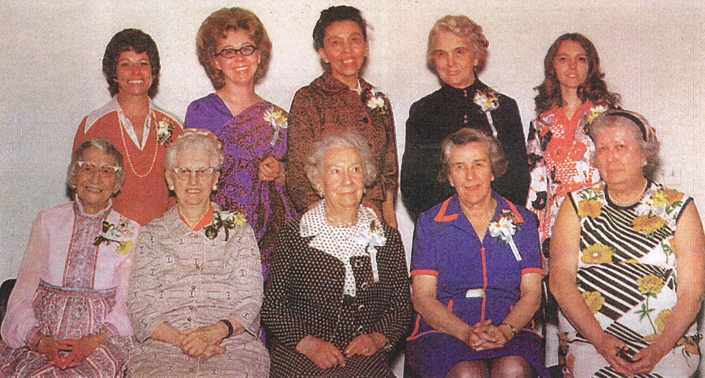 A group of ten women, most of them elderly, dressed in colorful vintage clothing, posing together in front of a plain wall.
