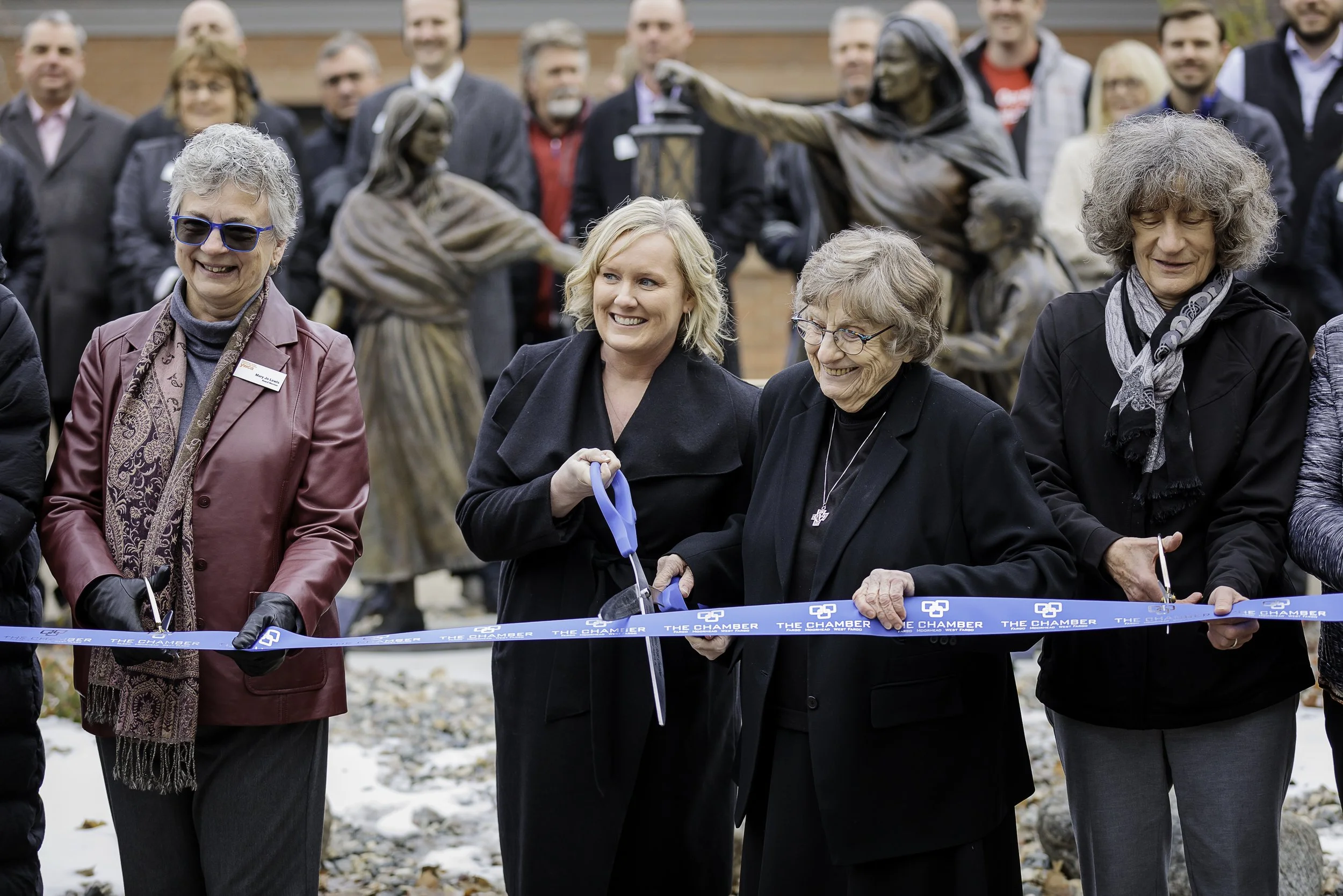 Women participate in a ribbon-cutting ceremony outdoors, with a crowd of onlookers and bronze statues of people in the background.