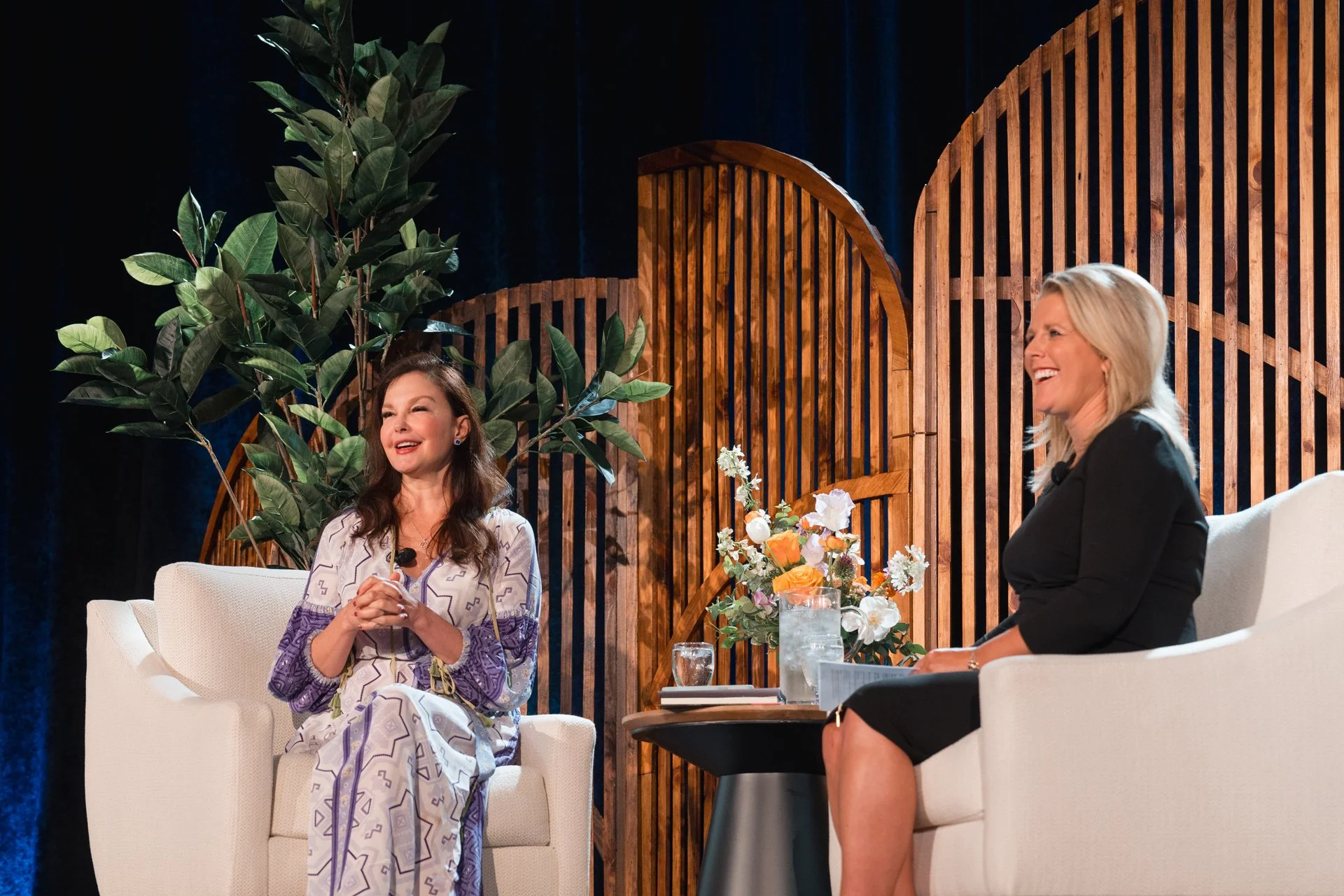 Ashley Judd and Kerstin Kealy sitting on white chairs in front of wood arch backdrop