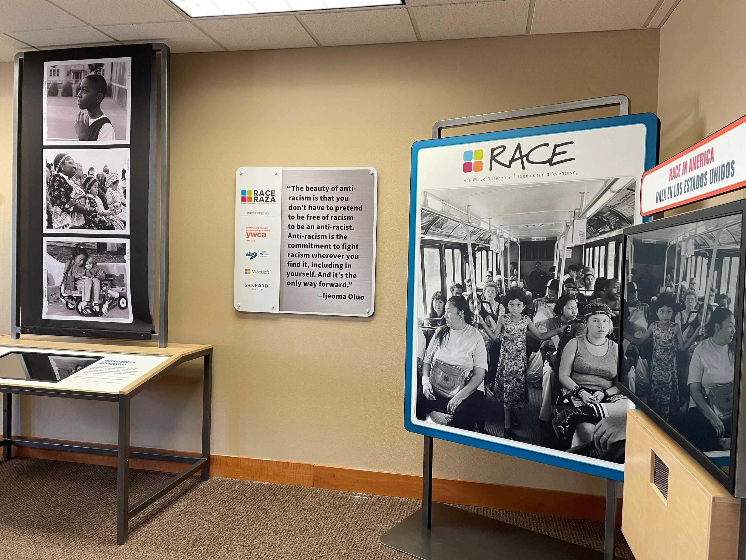 Indoor exhibition display with black-and-white photos of diverse children and families, a quote about anti-racism, and a poster titled 'RACE' showing people on a bus.