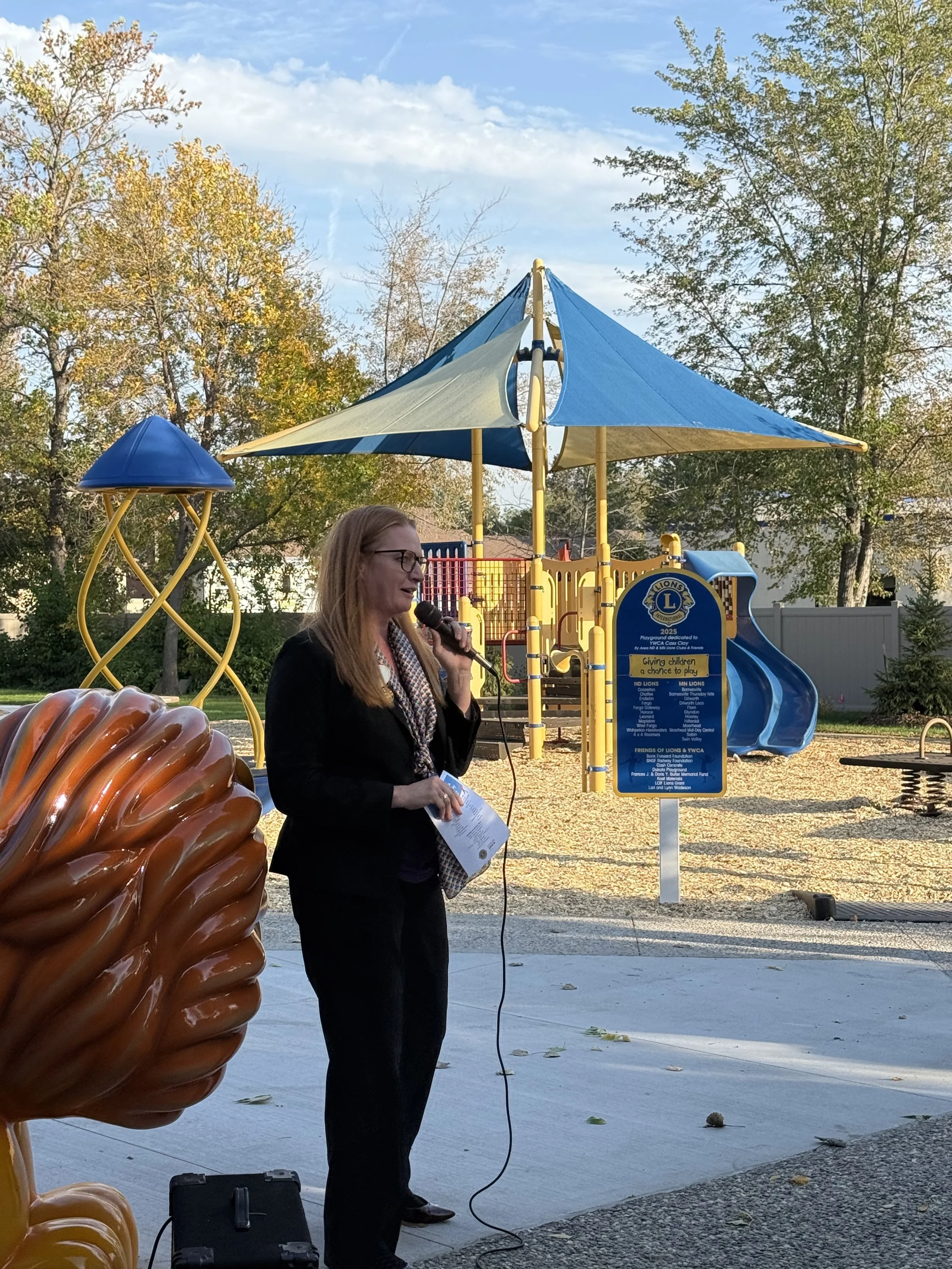 Woman speaking into microphone in front of blue and yellow playground
