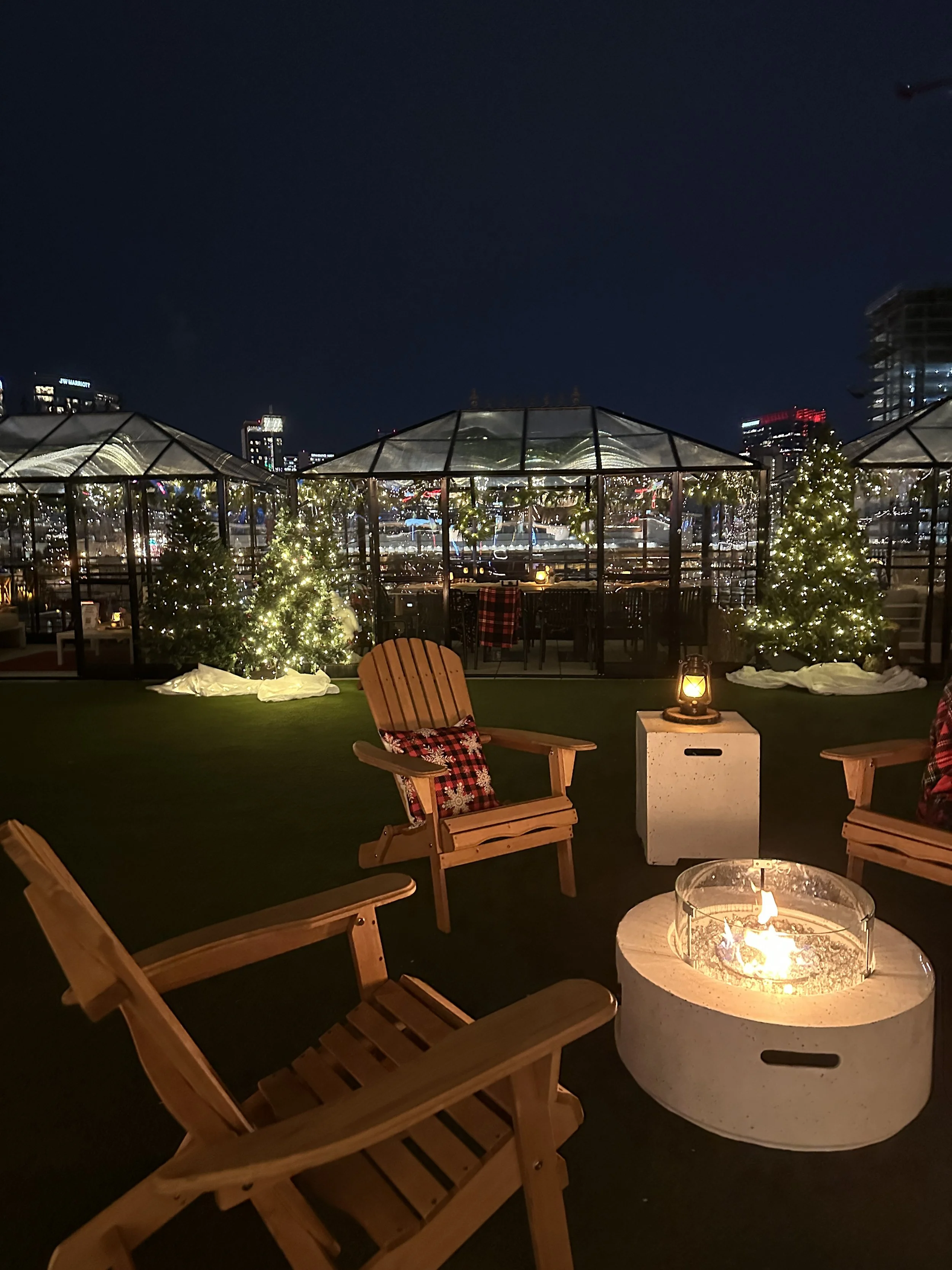 Nighttime rooftop patio decorated for Christmas with small illuminated Christmas trees, a fire pit, chairs with festive pillows, lantern lighting, and city skyline in the background.