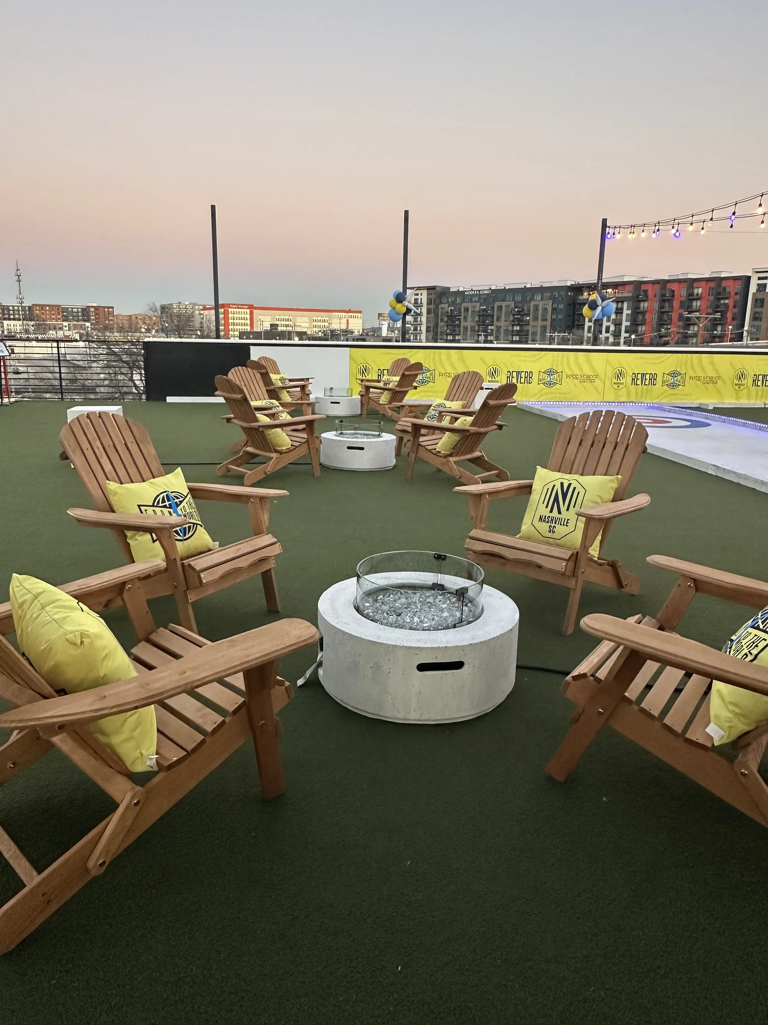 Rooftop seating area with wooden Adirondack chairs, yellow Nashville SC pillows, and fire pits, with city buildings and string lights in the background at sunset.
