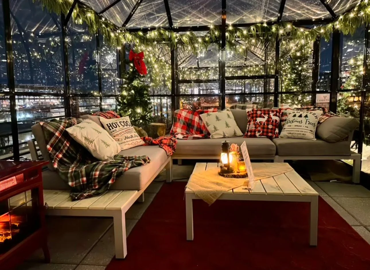 Cozy indoor holiday scene with plaid pillows, Christmas trees with lights, and string lights on the ceiling, overlooking a city at night.