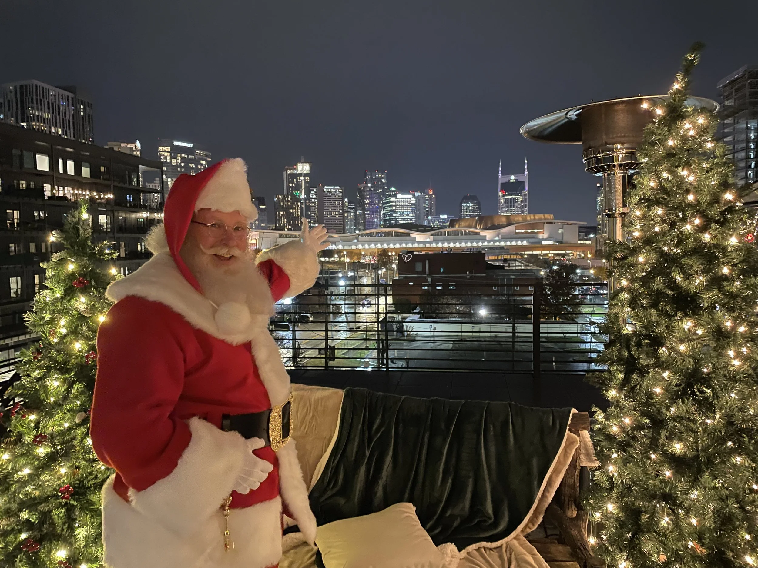 Santa Claus on a balcony with city skyline at night, decorated with Christmas trees and lights.