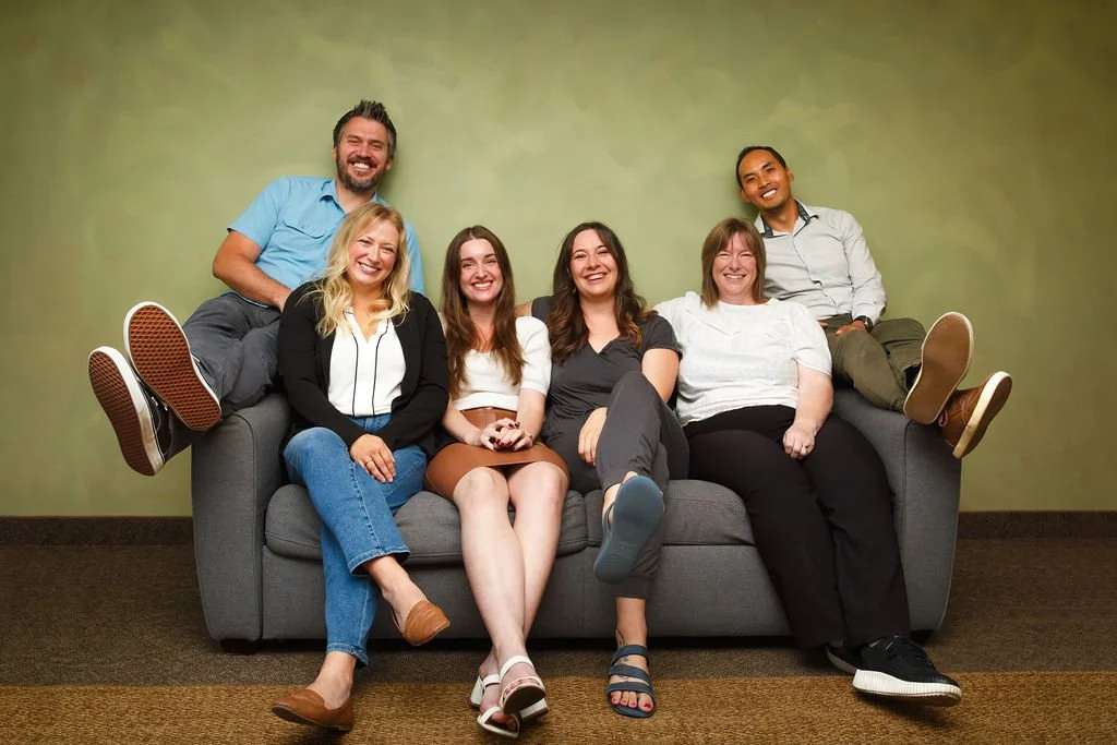 A group of six people sitting on a gray sofa against a green wall, smiling and relaxed. Four women are sitting in the front, and two men are sitting behind them on the armrest.