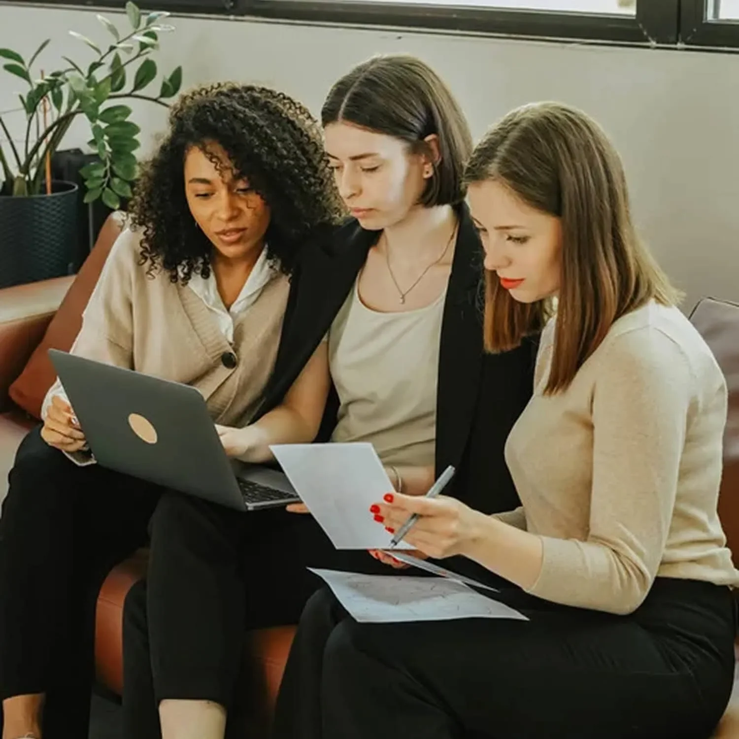 Three women sitting on bench reviewing work together with laptop and notes