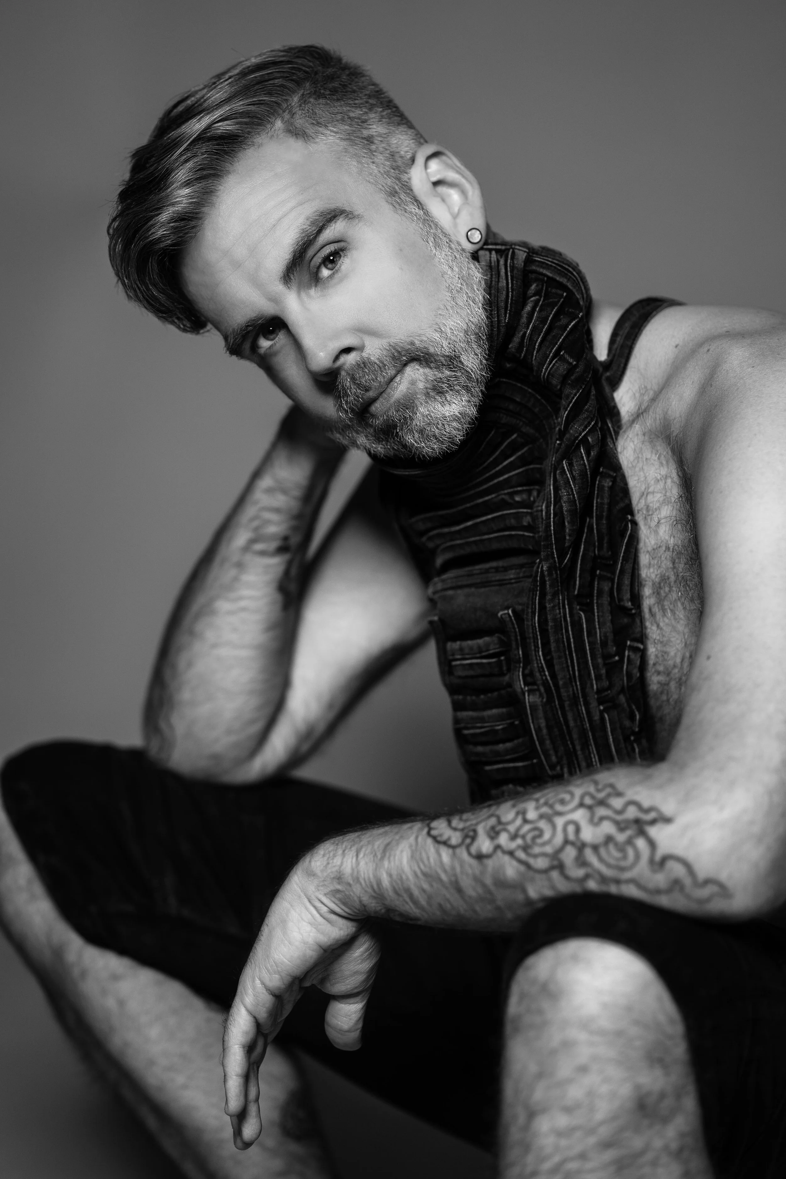 Black and white portrait of a bearded man with styled hair, wearing a sleeveless vest and shorts, sitting with one arm resting on his knee. He has tattoos on his arm and a small earring in his left ear, with a neutral background.