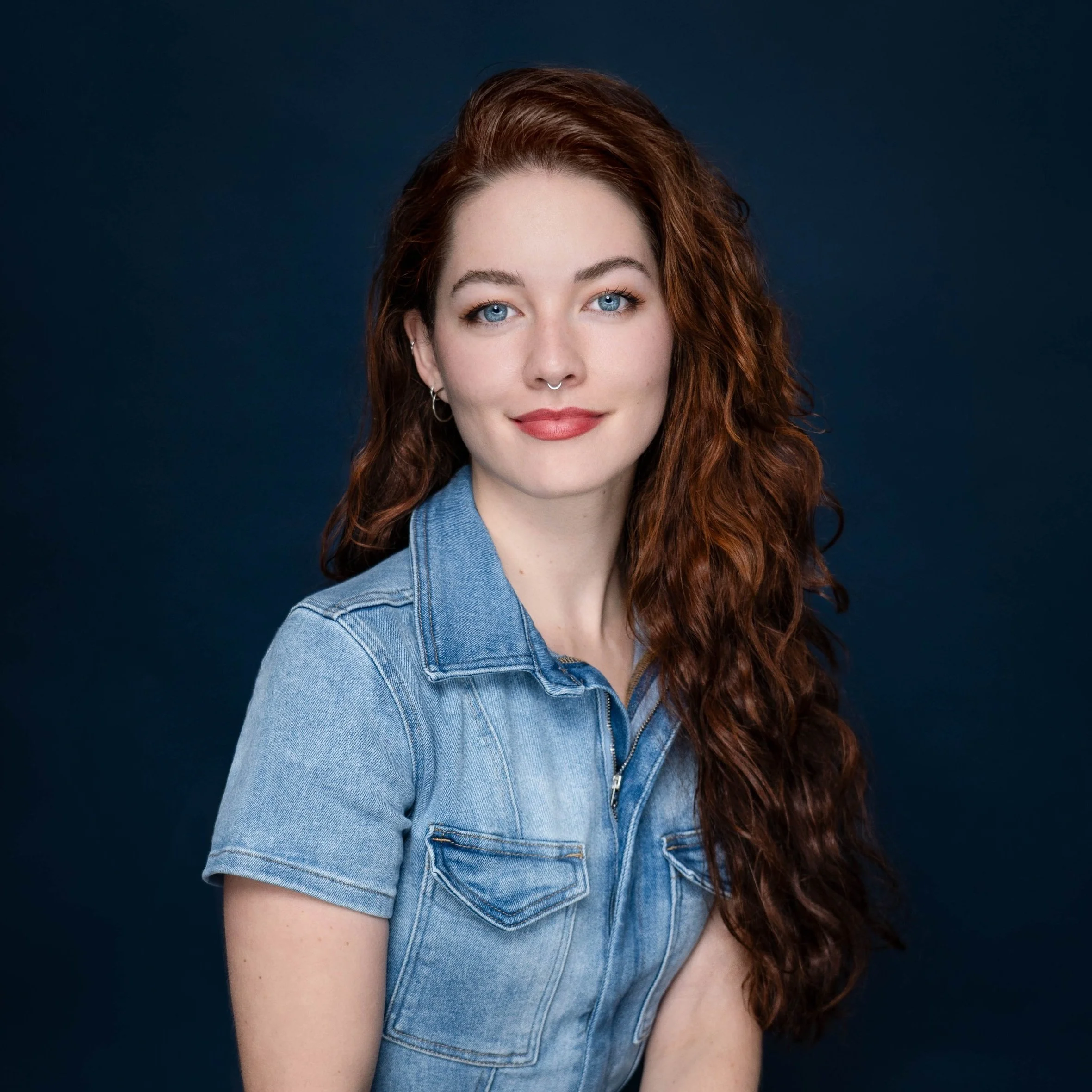 A young woman with long curly red hair, blue eyes, and a septum piercing, wearing a light blue denim short-sleeve shirt, smiling softly against a dark background.
