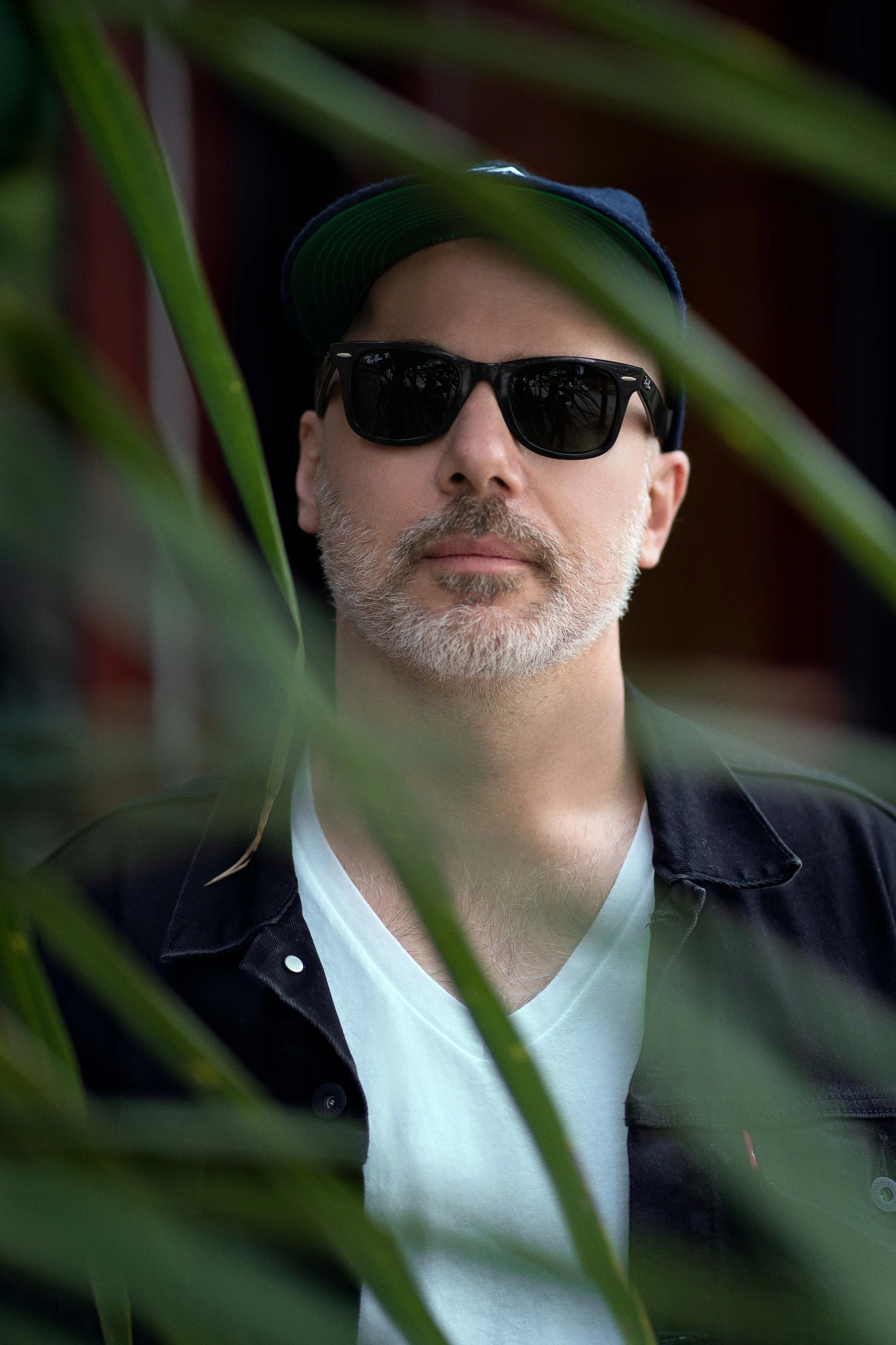 A man with a gray beard wearing sunglasses, a blue cap, a black jacket, and a white shirt, partially obscured by green leaves.