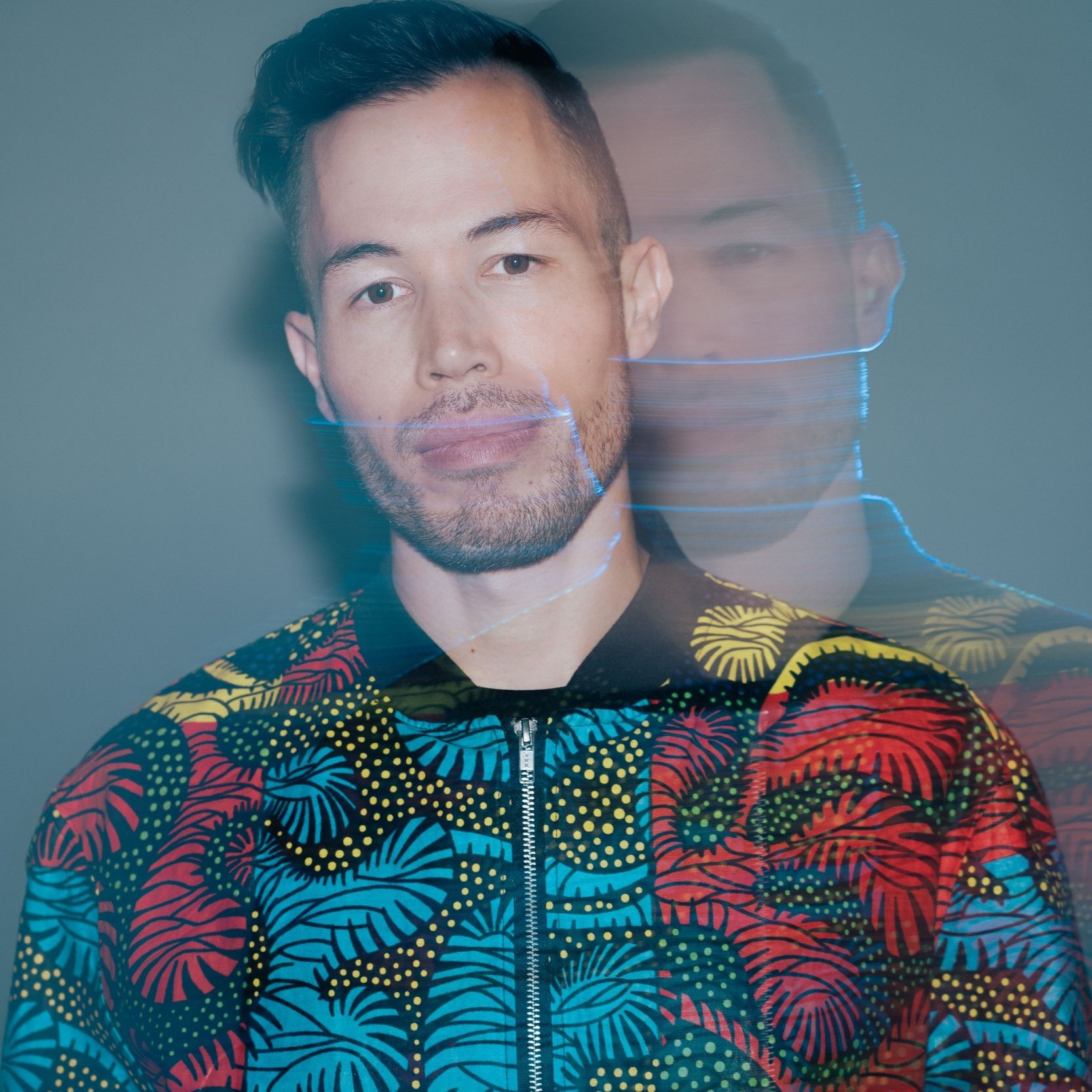 Portrait of a man with short dark hair and a beard wearing a colorful patterned jacket, with a reflection creating a double exposure effect.