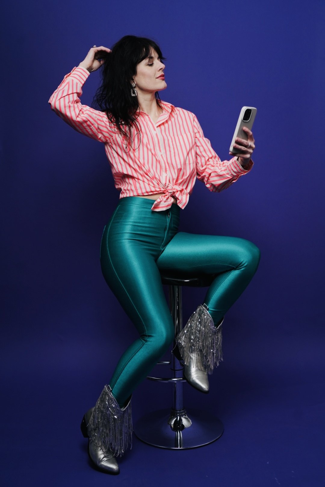 A woman with black hair wearing a pink and white striped tied shirt, teal shiny leggings, and silver fringe boots, sitting on a stool against a dark blue background, looking at her smartphone.