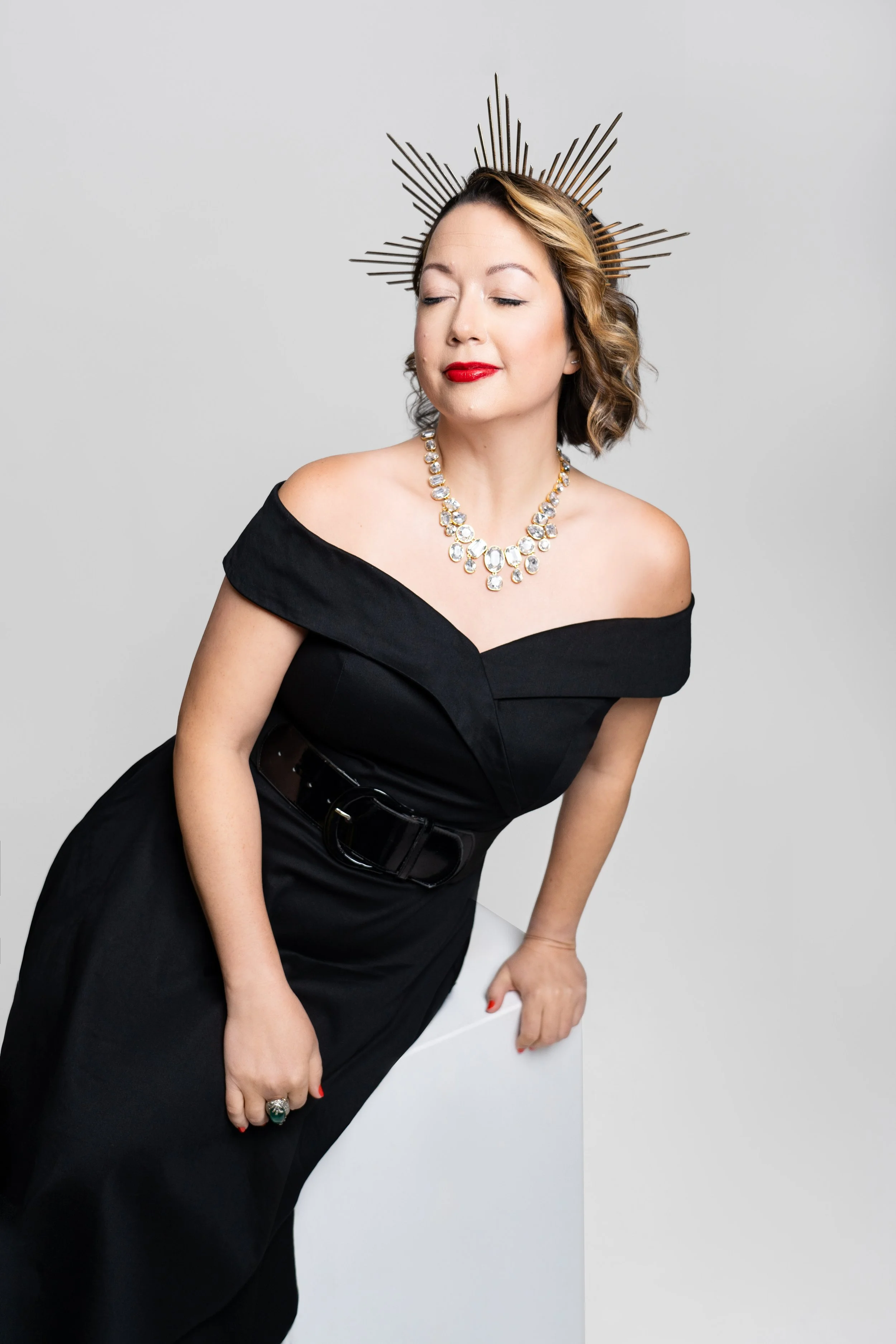 A woman with short curly hair wearing a black off-shoulder dress, a large diamond necklace, and a starburst headpiece, poses against a plain background.